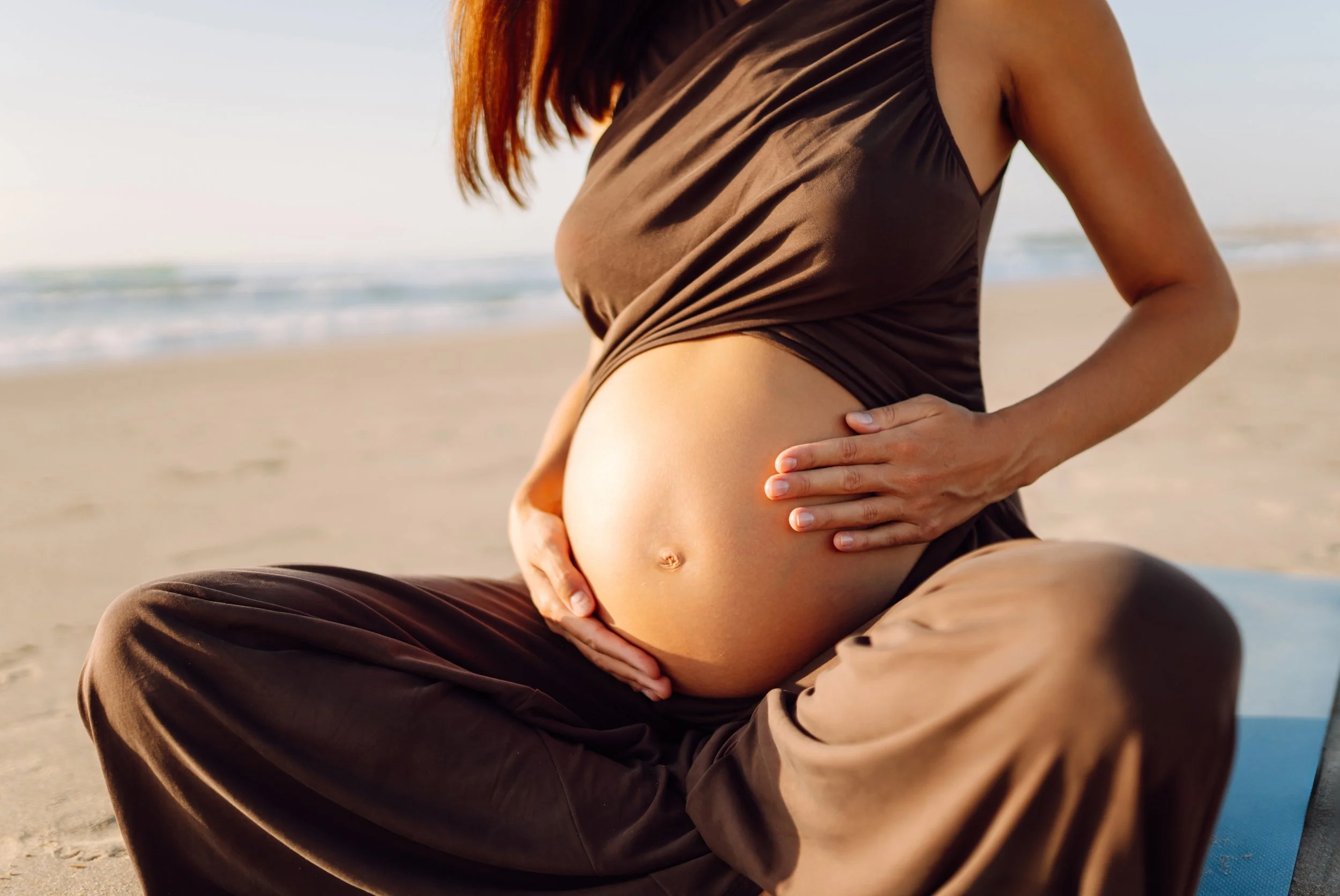 Pregnant woman sitting on the beach with her hand on her belly, wearing a brown dress.