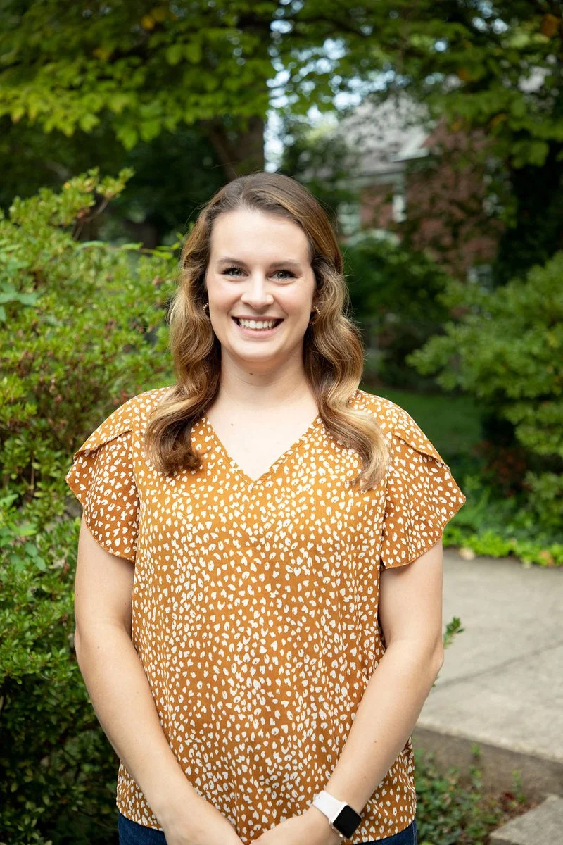 Young woman with wavy brown hair smiling outdoors, wearing a mustard yellow blouse with white pattern, standing in front of green trees and bushes.