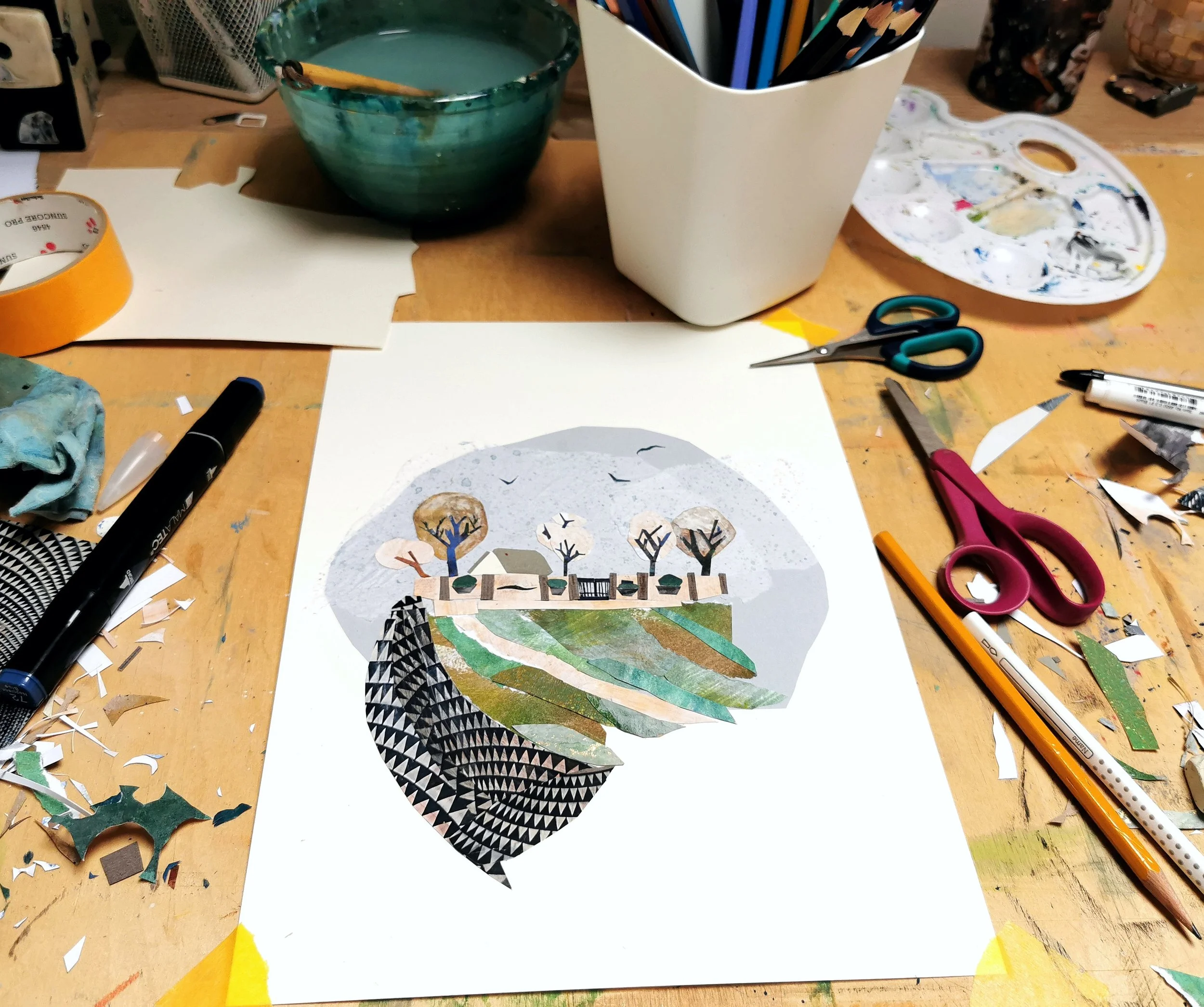 A collage artwork of a landscape scene with trees, a house, and a path, created with various textured paper on white paper, placed on a cluttered desk with scissors, markers, tape, brushes, and art supplies.