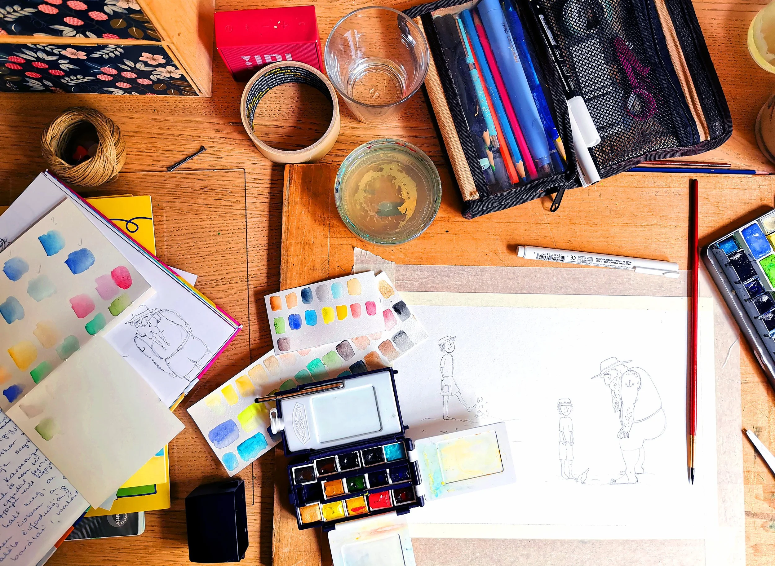 Art supplies on a wooden table, including watercolor paints, swatches, sketches, a water container, colored pencils, and a pencil drawing of characters.