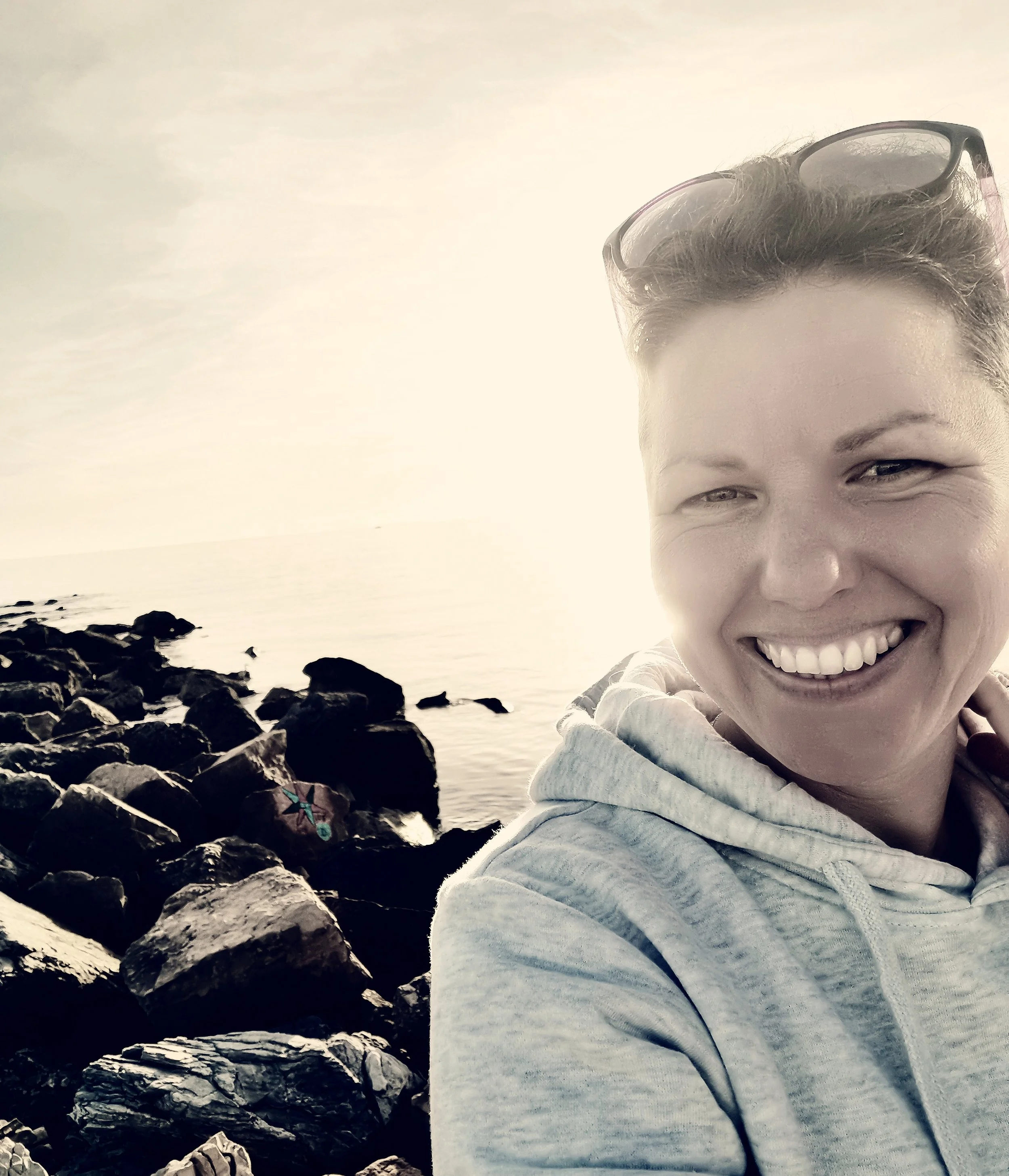 A woman smiling at the camera outdoors near a rocky shoreline and calm water, with the sun bright in the background, wearing sunglasses on her head and a light hoodie.