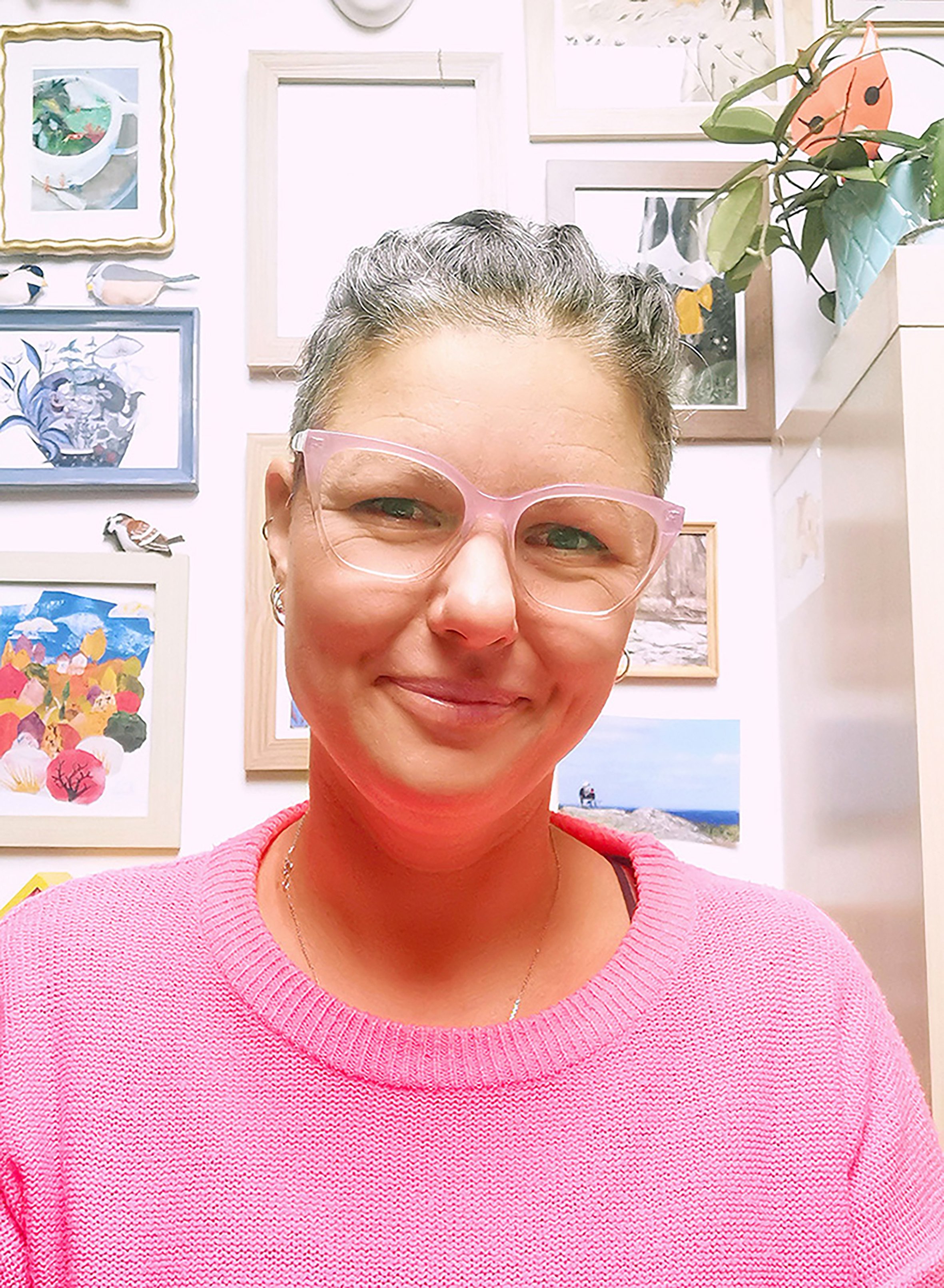 A woman wearing pink glasses and a pink sweater, smiling in front of a wall decorated with art and photographs.