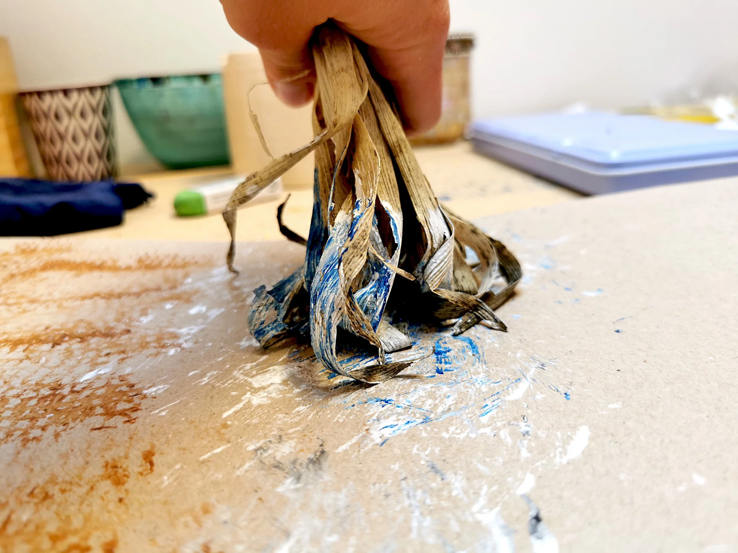 A hand holding a bunch of burned, twisted paper strips on a cluttered table with various objects in the background.