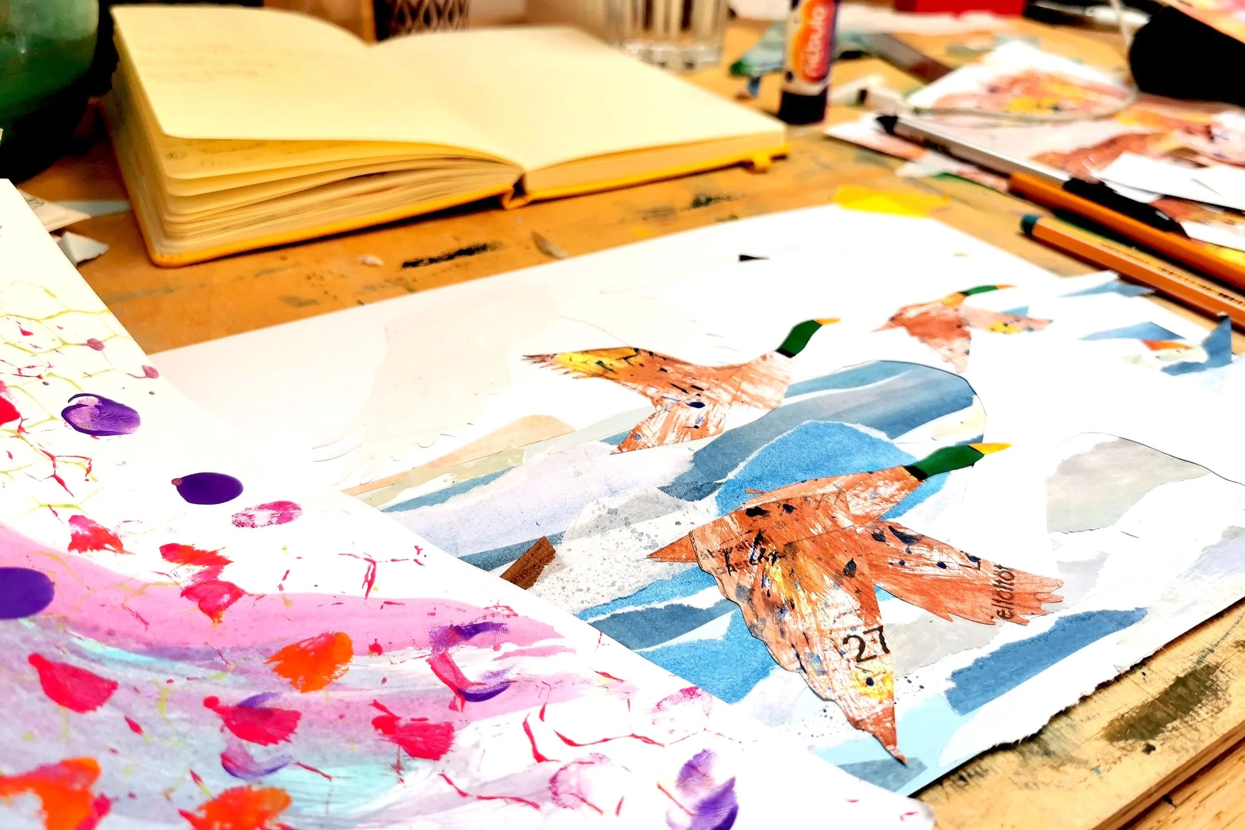 Colorful collage artwork of geese flying over mountains on paper, surrounded by art supplies on a wooden table, including paint, markers, and a sketchbook.