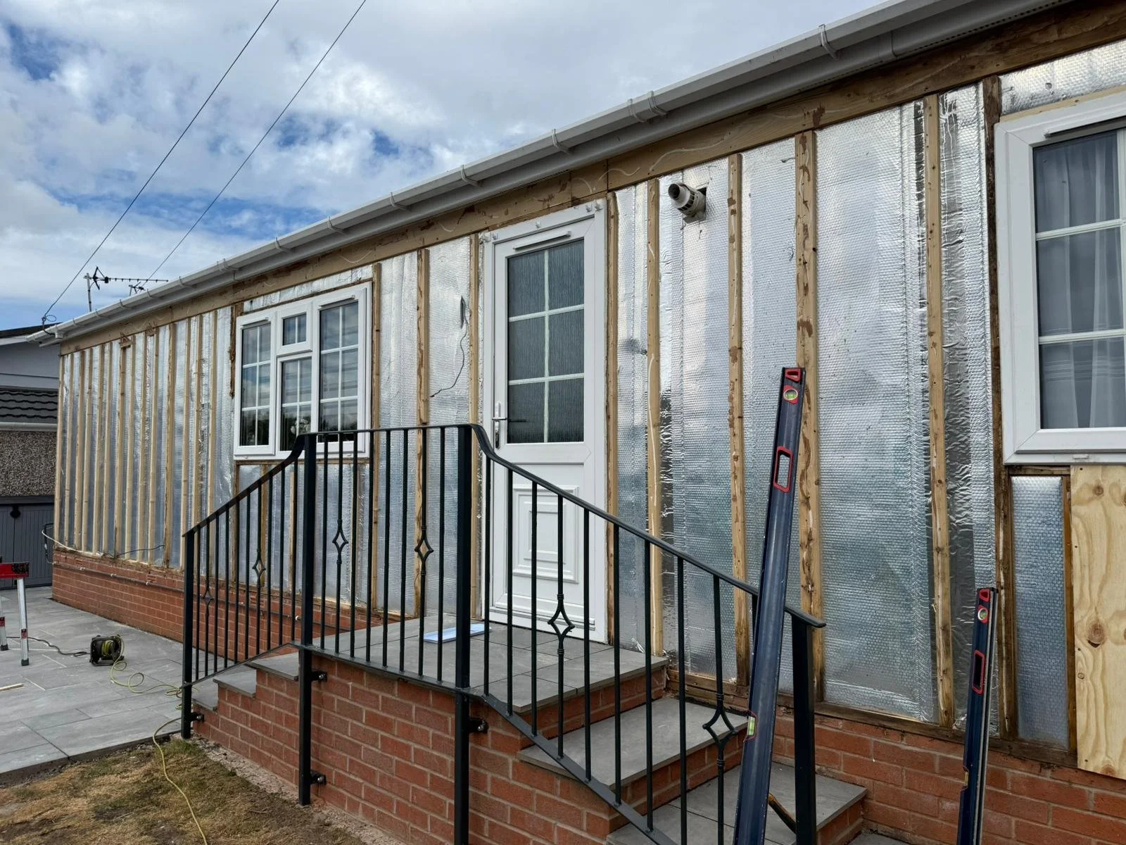 Park home exterior under renovation, with exterior walls partially covered with insulation and wooden framing, a white door with window panels, three windows, a metal railing staircase, and construction tools including level and electrical cord.