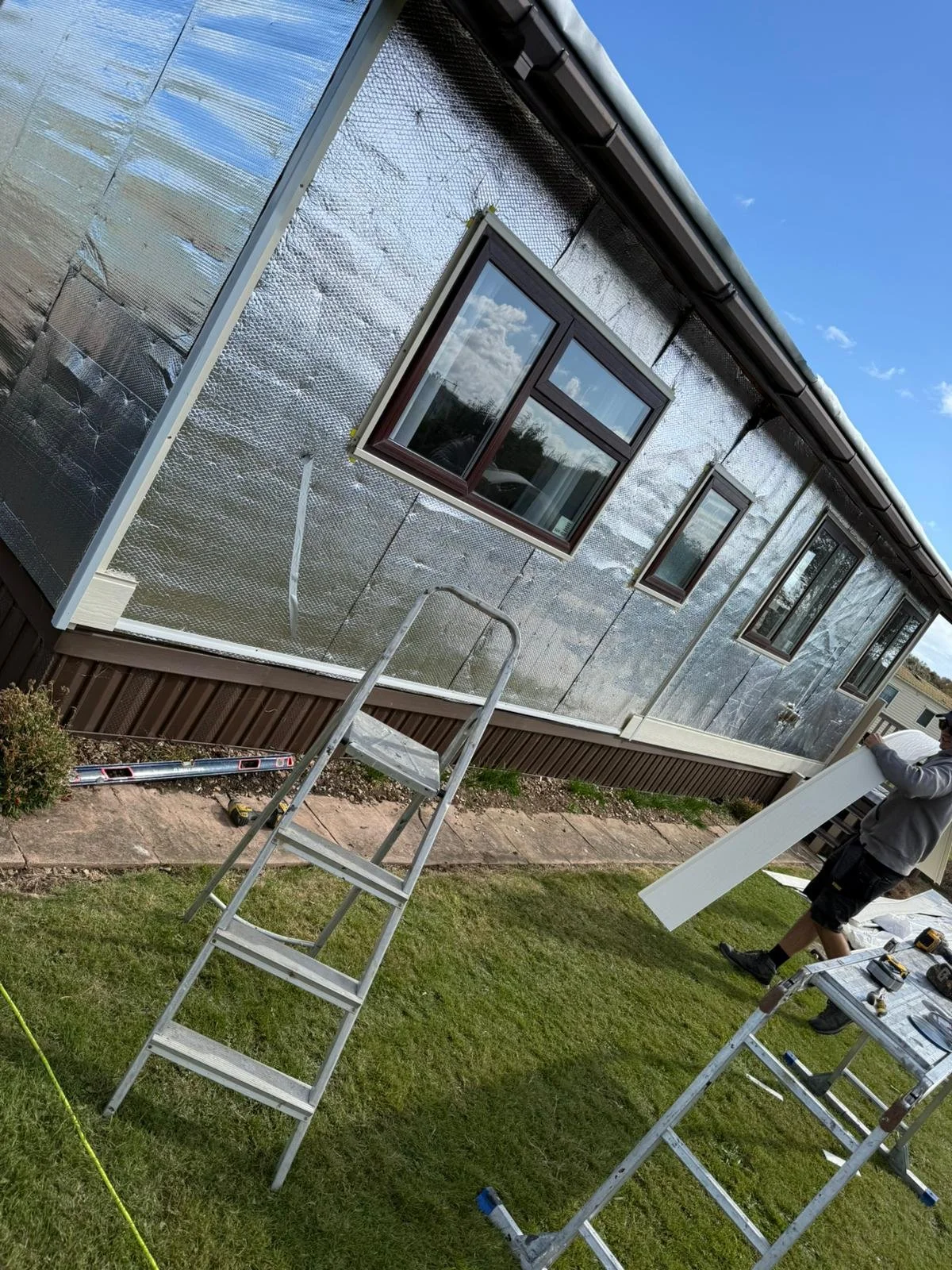 Construction worker installing a white siding panel on the exterior of a park home. The park home has three windows and reflective insulation.