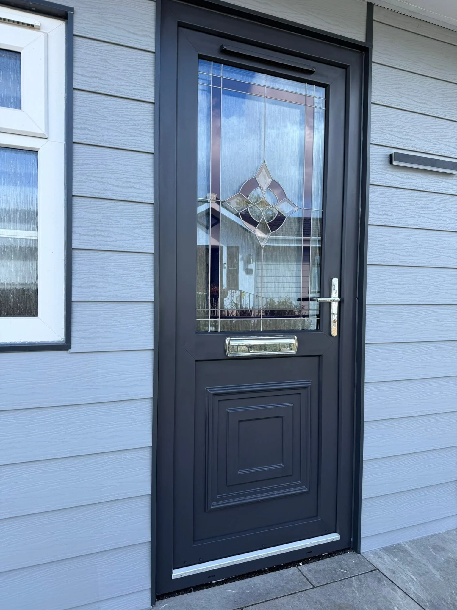 Park home front door with glass panel and decorative stained glass design, black frame, silver handle, set in a house with light grey horizontal cladding, and window to the left.