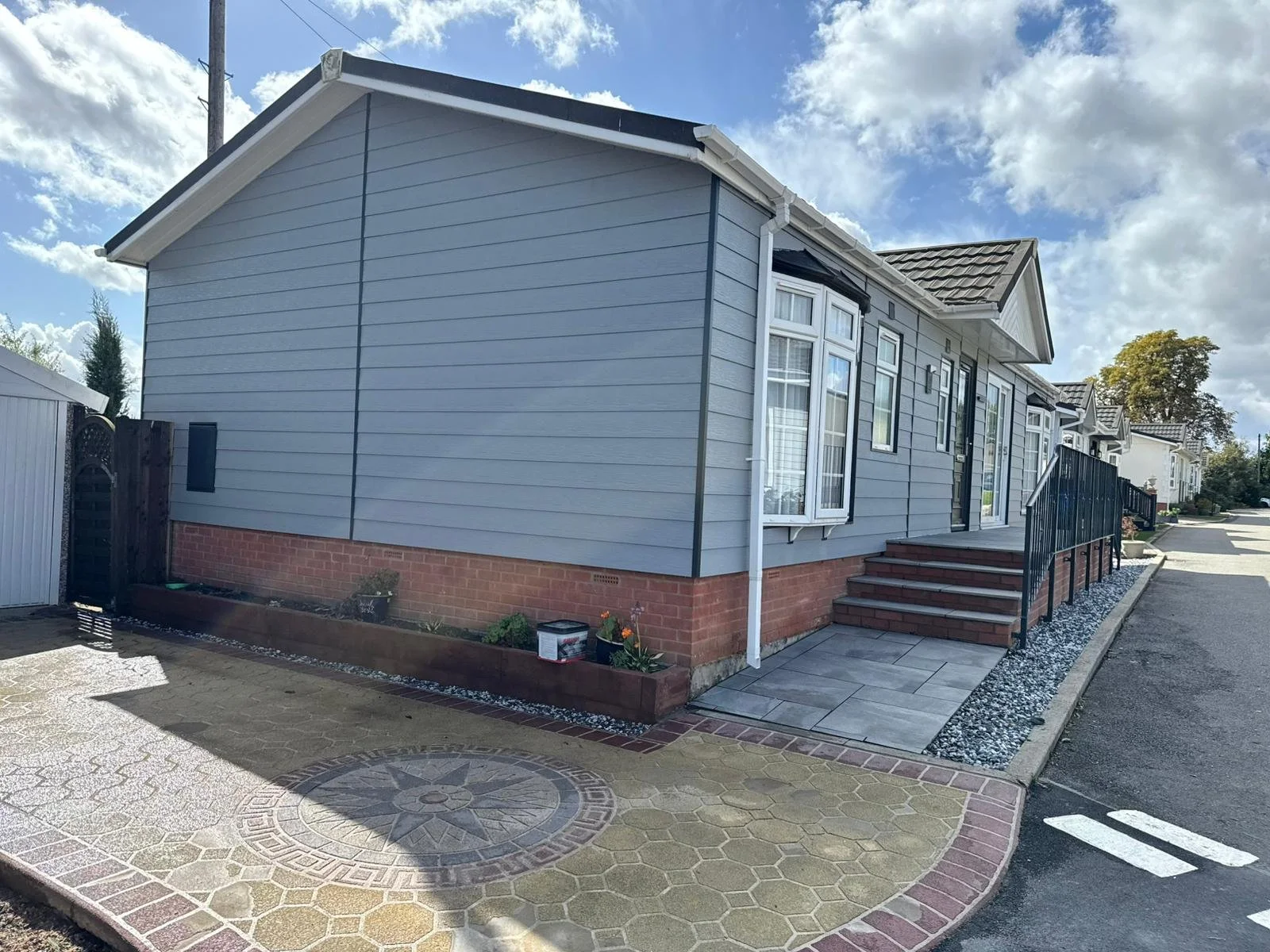 Side view of a modern park home with light grey cladding, black roof, red brick foundation, stairs leading to a door, and a small garden bed with some plants and potted flowers outside.