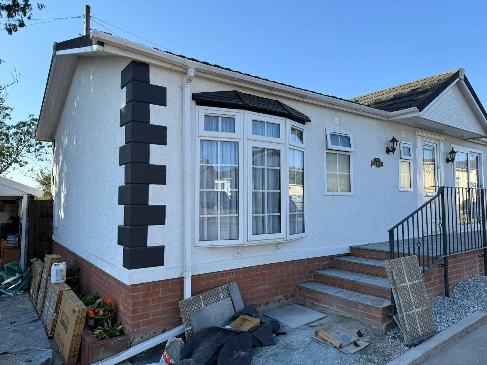 A white park home with black decorative bricks on the corner, large windows, and a small staircase leading to the front door, under a clear blue sky.