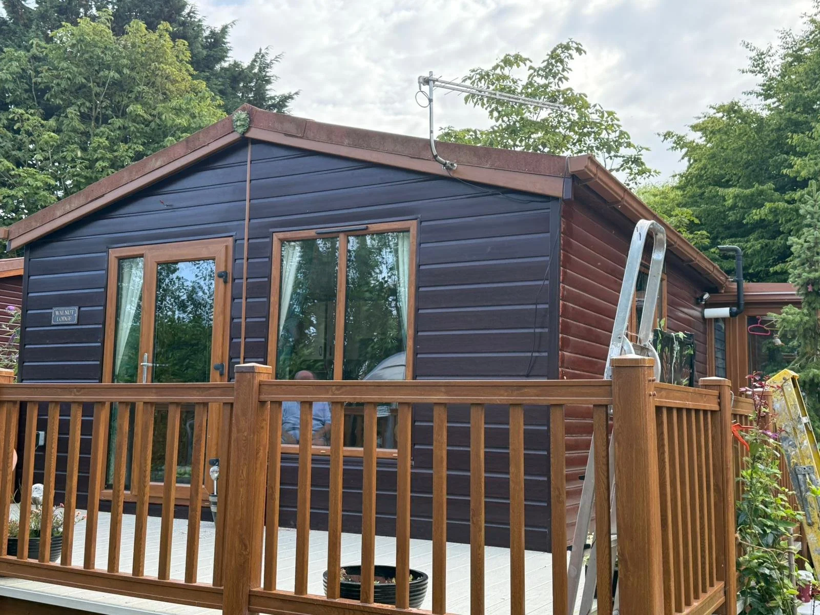 A small wooden park home with dark  brown exterior cladding, glass sliding door, and a wooden deck surrounded by a wooden railing, with trees and cloudy sky in the background.