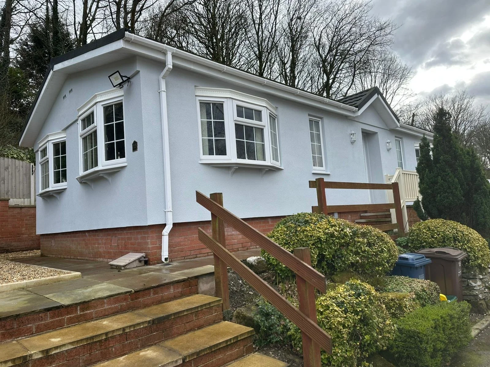 A white park home with bay windows, brick foundation, and a small porch with a wooden staircase and railing. There are bushes and a garbage bin in the front yard, with trees and a cloudy sky in the background.
