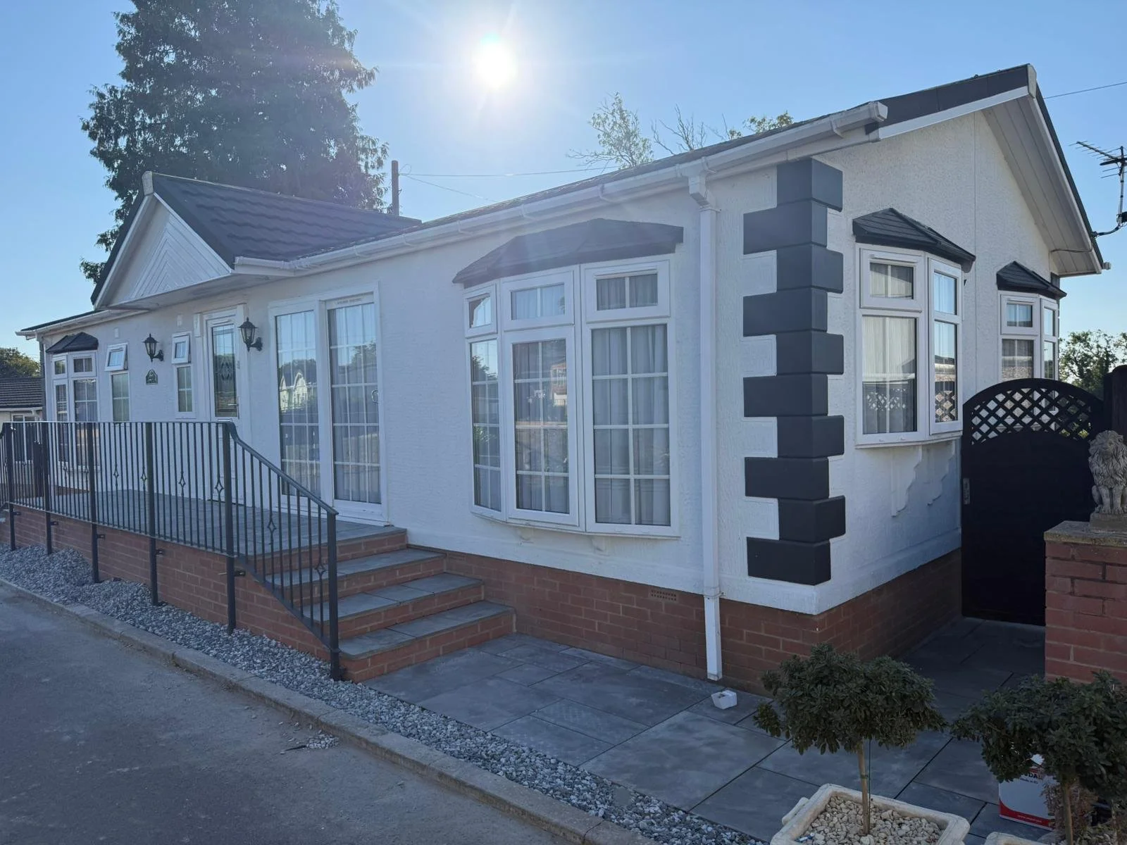 Exterior of a white park home with a black gate and steps leading to a door. The house has multiple windows with white frames and shutters, a dark grey corner embellishment, a brick base, and a tiled roof. The sun is bright in the sky.