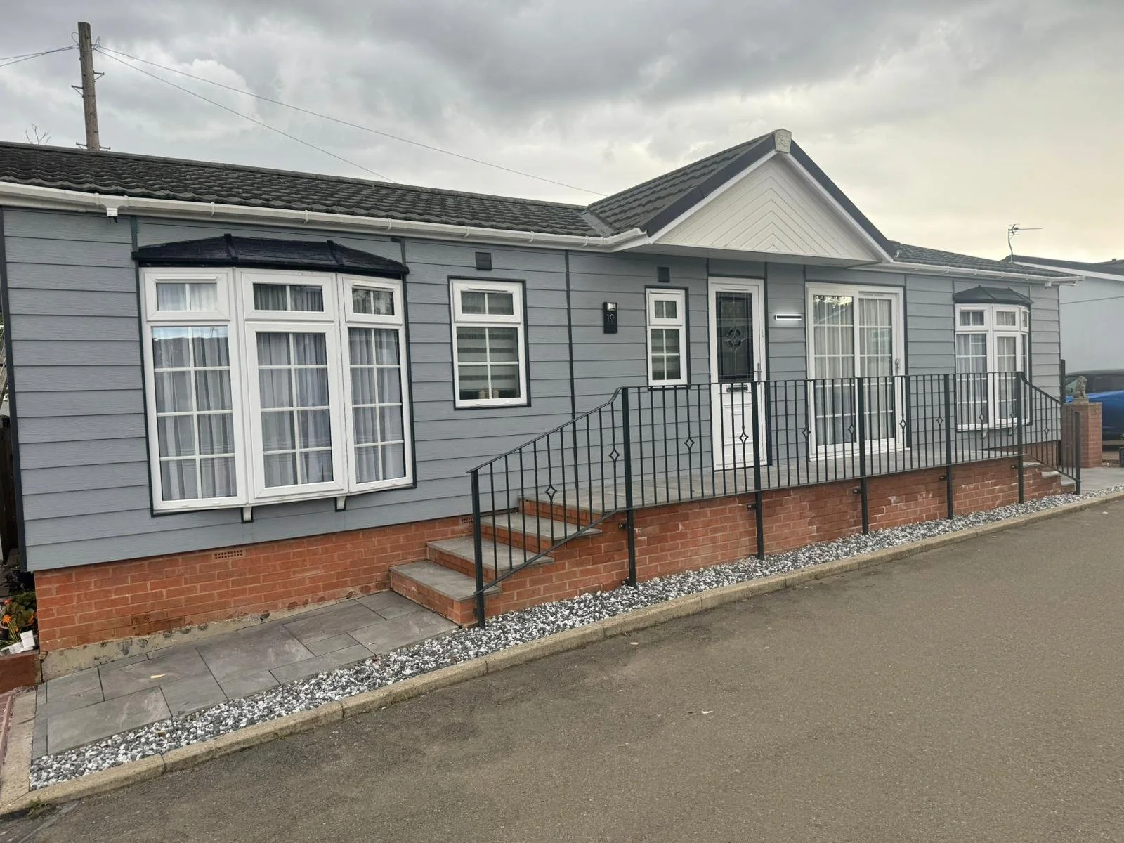 A park home with grey cladding, white trim, and a brick foundation. It features bay windows with white curtains, a small staircase leading to the front door, and a black metal railing.