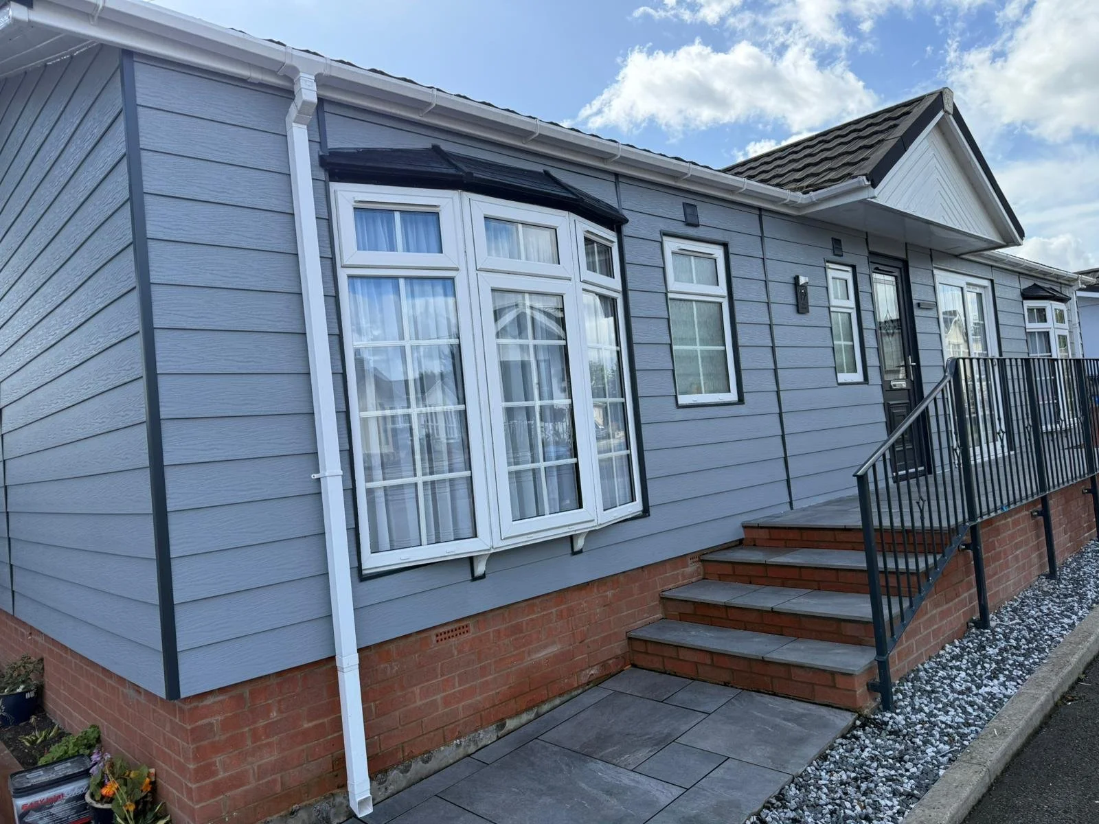 Exterior of a modern park home with grey cladding, a bay window, and a small set of wooden stairs with a black metal railing leading to the front door on a partly cloudy day.
