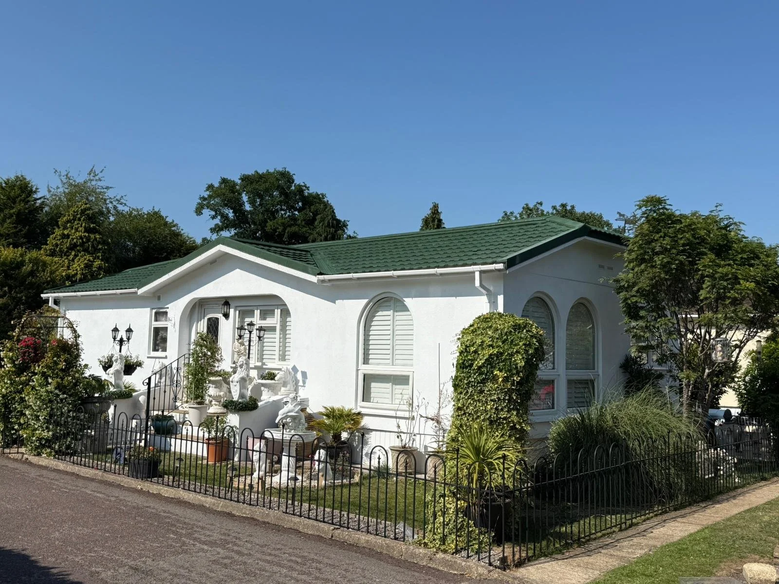 A white park home with a green roof, surrounded by a metal fence, with various plants and garden ornaments in the front yard under a clear blue sky.
