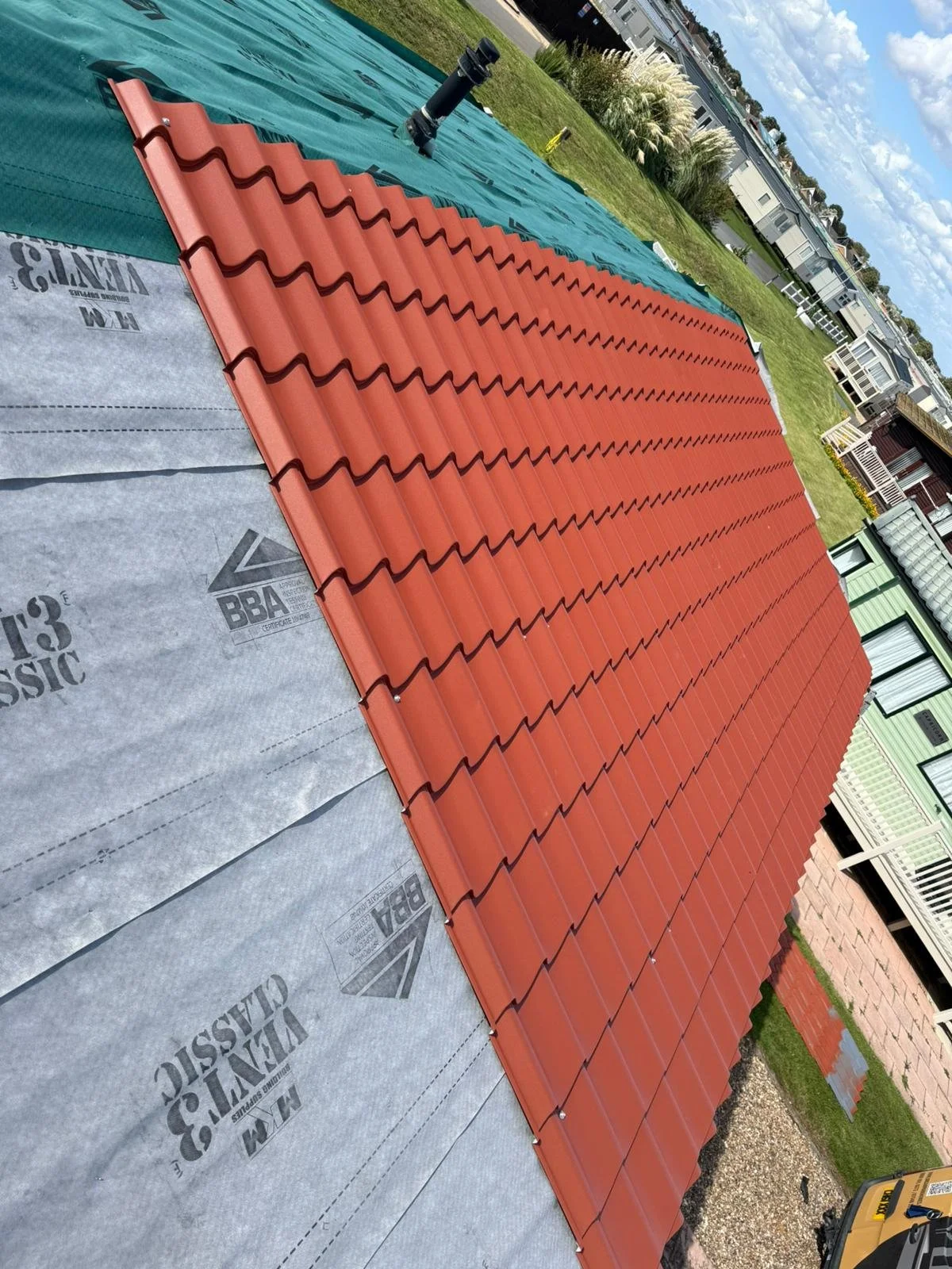 Photograph of a park home roof under construction with red clay tiles installed, grey underlayment visible at the bottom, and residential buildings and a partly cloudy sky in the background.