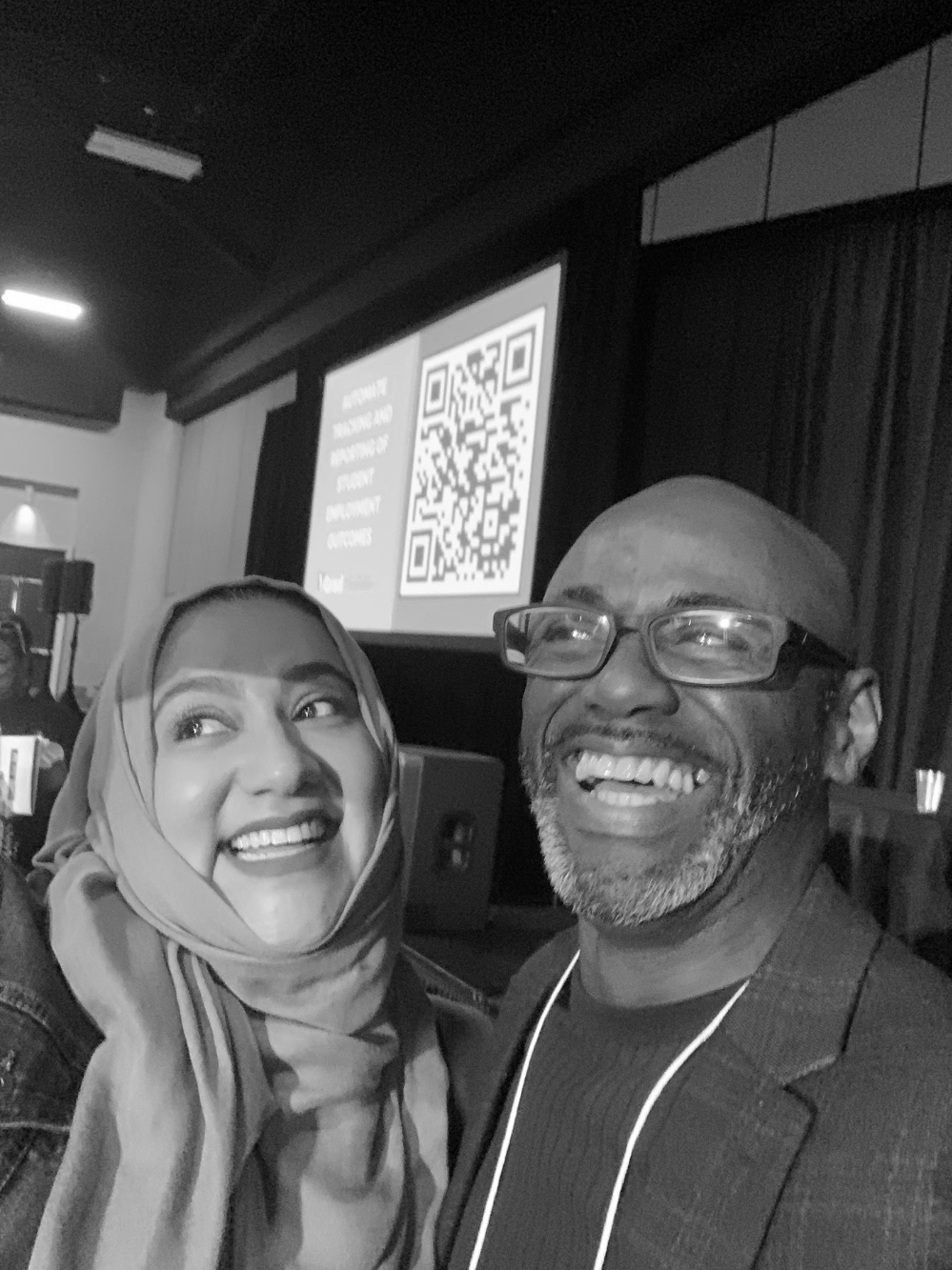 A woman and a man smiling and posing for a selfie at an indoor event with a large screen and a QR code in the background.