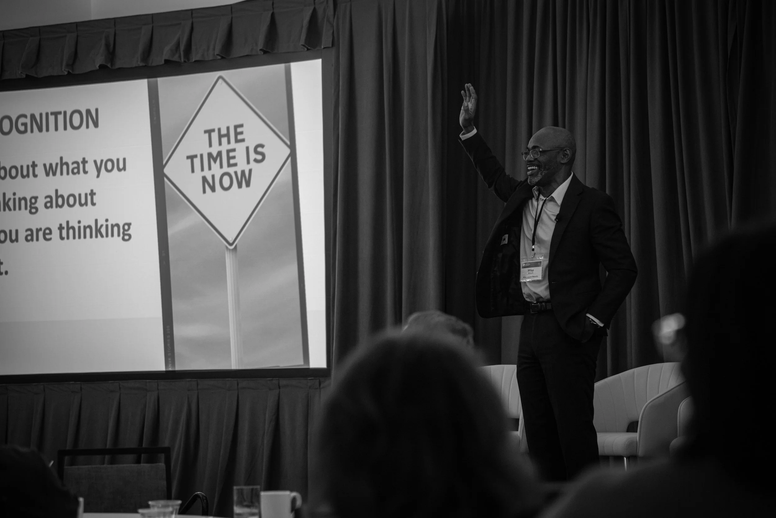 A man in a suit and glasses giving a presentation on stage, with a slide that reads 'The Time Is Now' and a message about thinking and talking.