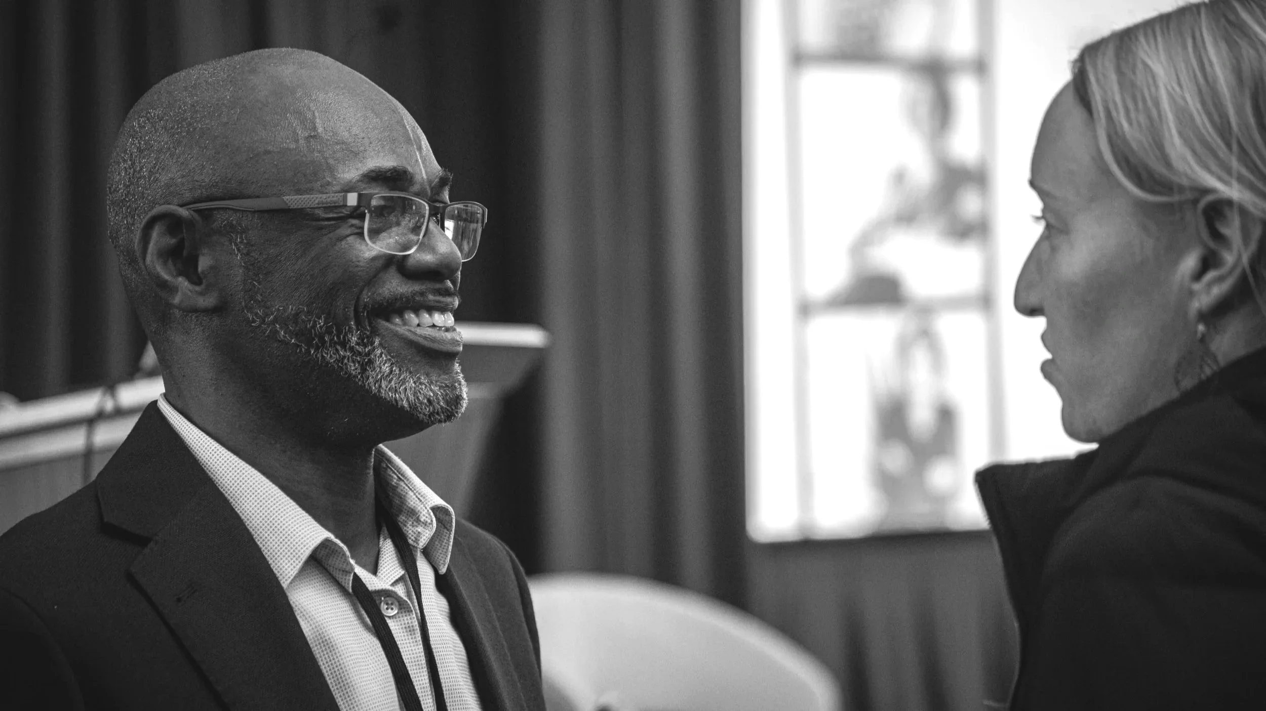 Two people talking in an indoor setting, one man smiling and the woman listening attentively, both in profile view.