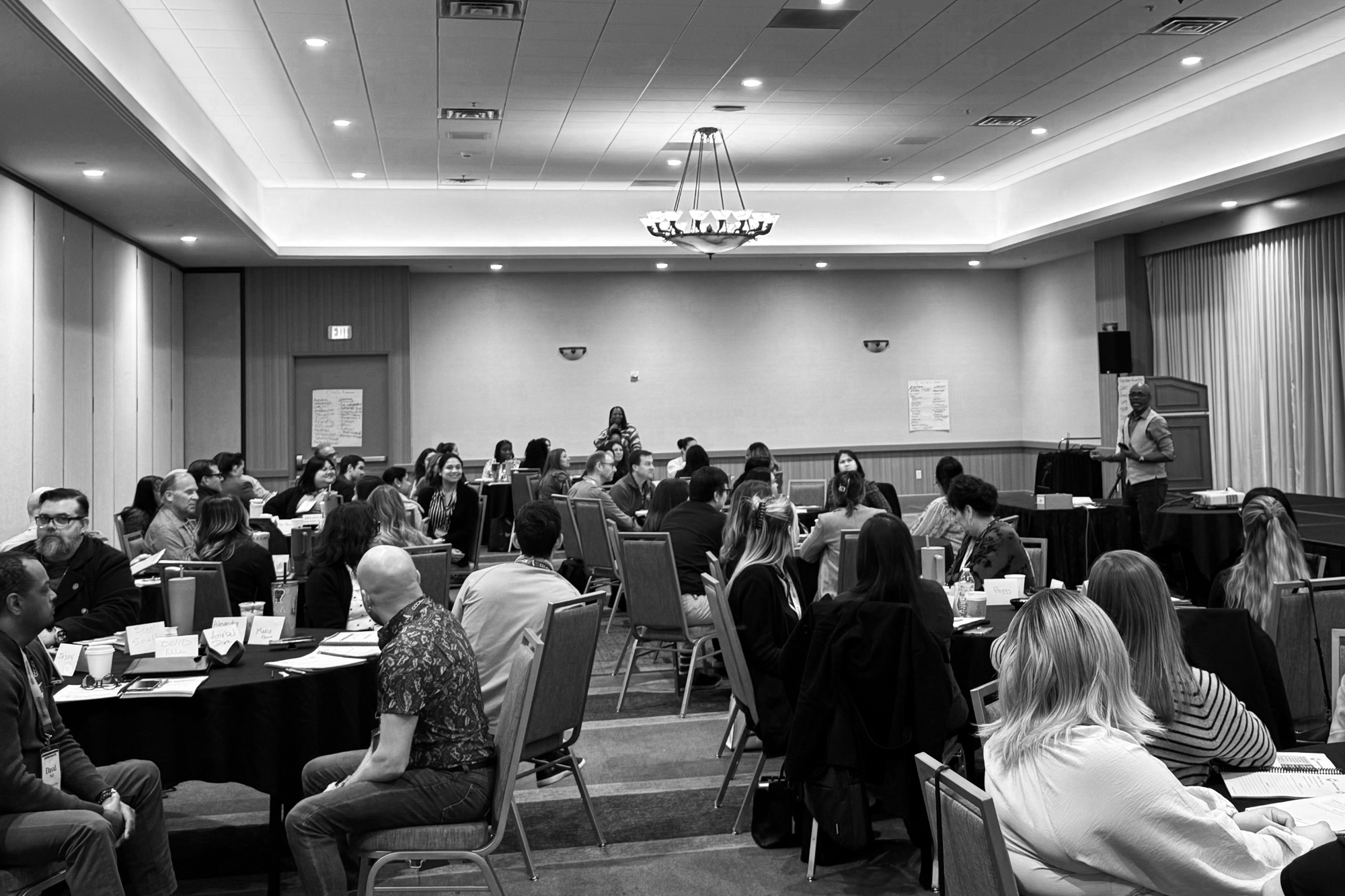 Black and white photo of a conference room filled with people seated at round tables, facing a stage where a man is standing with a microphone, with a woman on the stage.