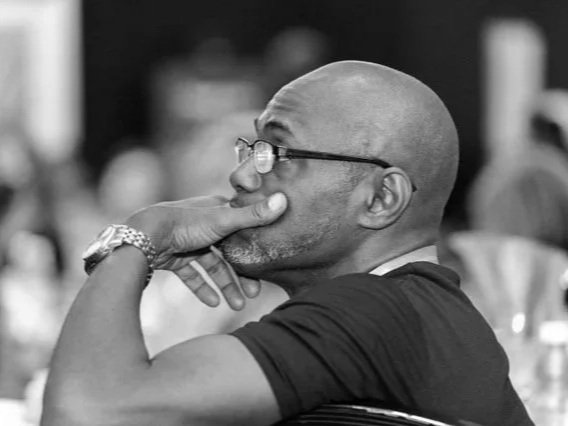 A bald man wearing glasses, resting his chin on his hand, in a candid moment, in black and white.
