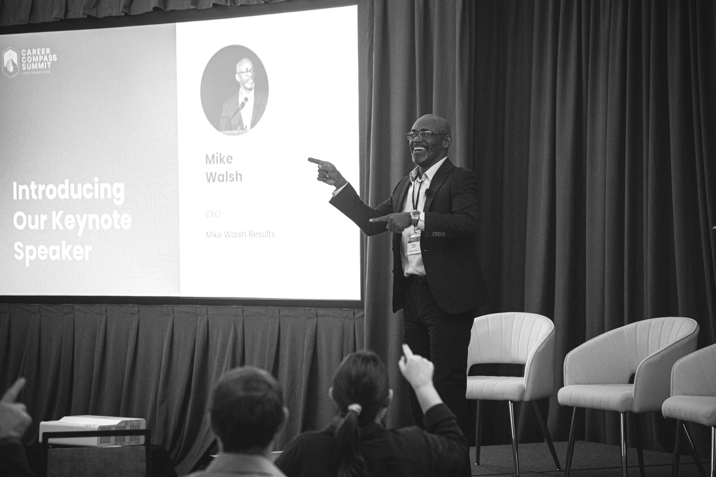 Man in a suit standing on stage, pointing at a projected presentation slide with his right hand, smiling. The slide shows his photo and introduces him as Mike Walsh, CEO.