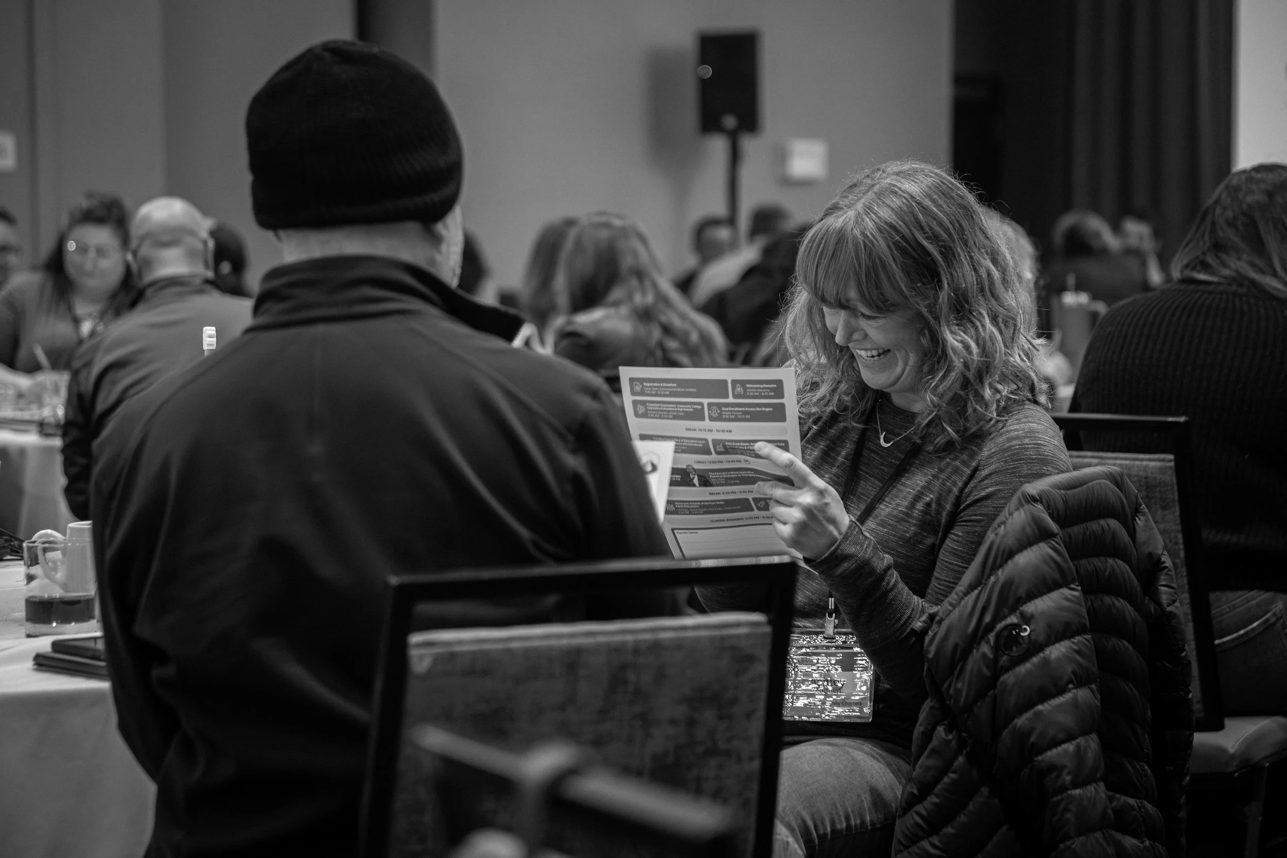 A woman with curly hair smiling and looking at a pamphlet while sitting in a conference room next to a man wearing a beanie. Other people are seated at tables in the background.