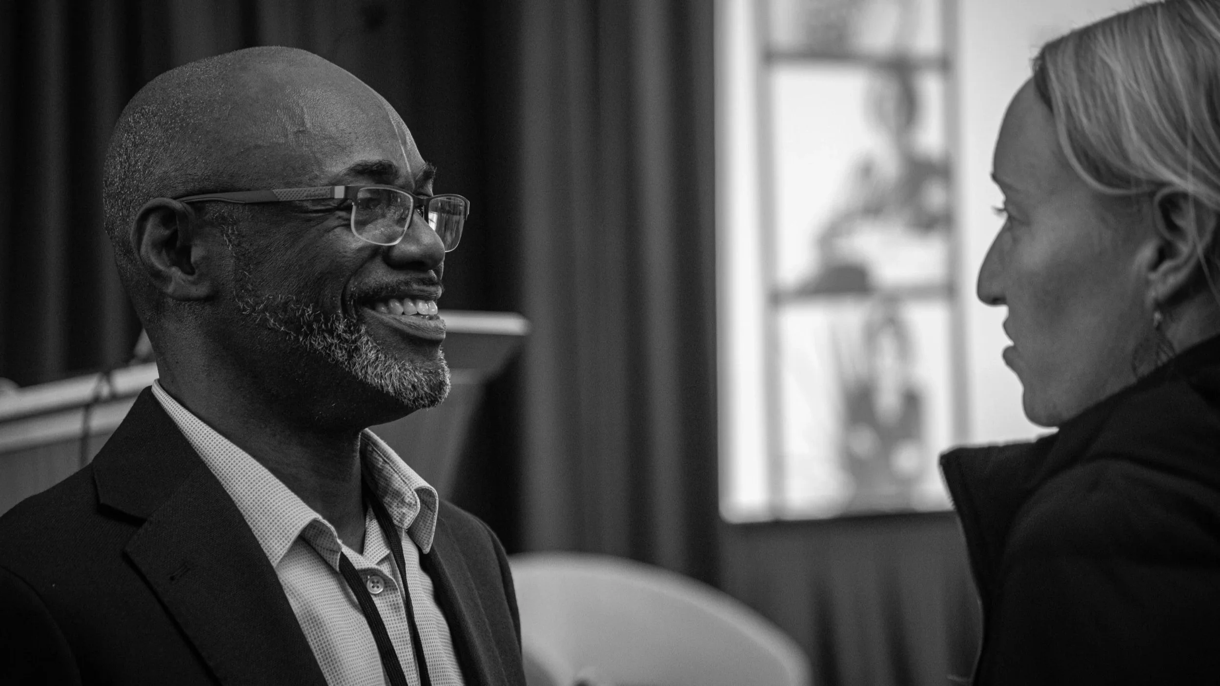 A Black man with glasses and a beard smiling and talking to a white woman with light hair, in a room with wooden paneling and a window with blurred figures in the background.