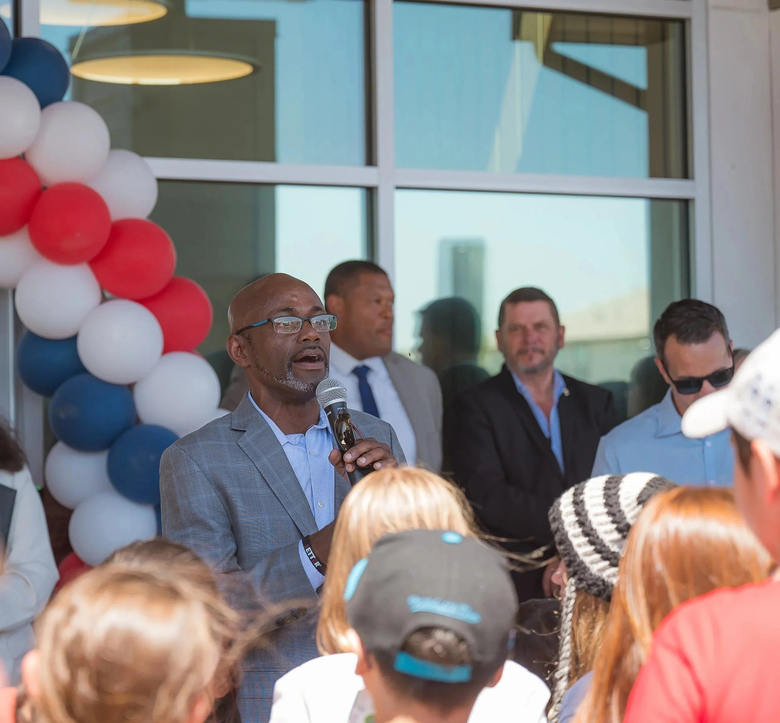 A man wearing glasses and a gray suit is speaking into a microphone at a crowd event outdoors, with a decorative balloon arch in red, white, and blue in the background. Several people are standing behind him, some listening attentively.
