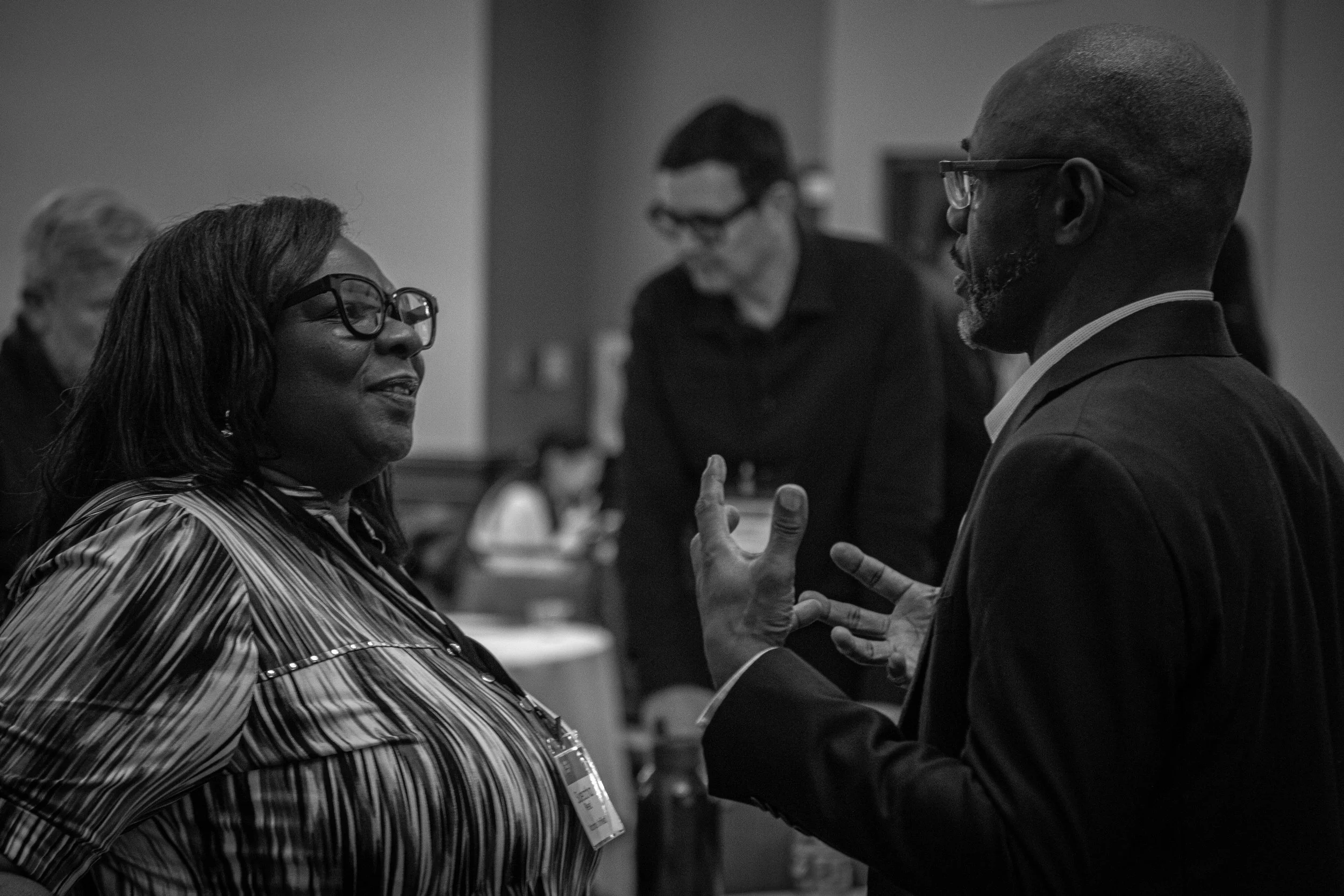 Two people engaged in conversation with smiling expressions, in a professional setting, with others in the background.
