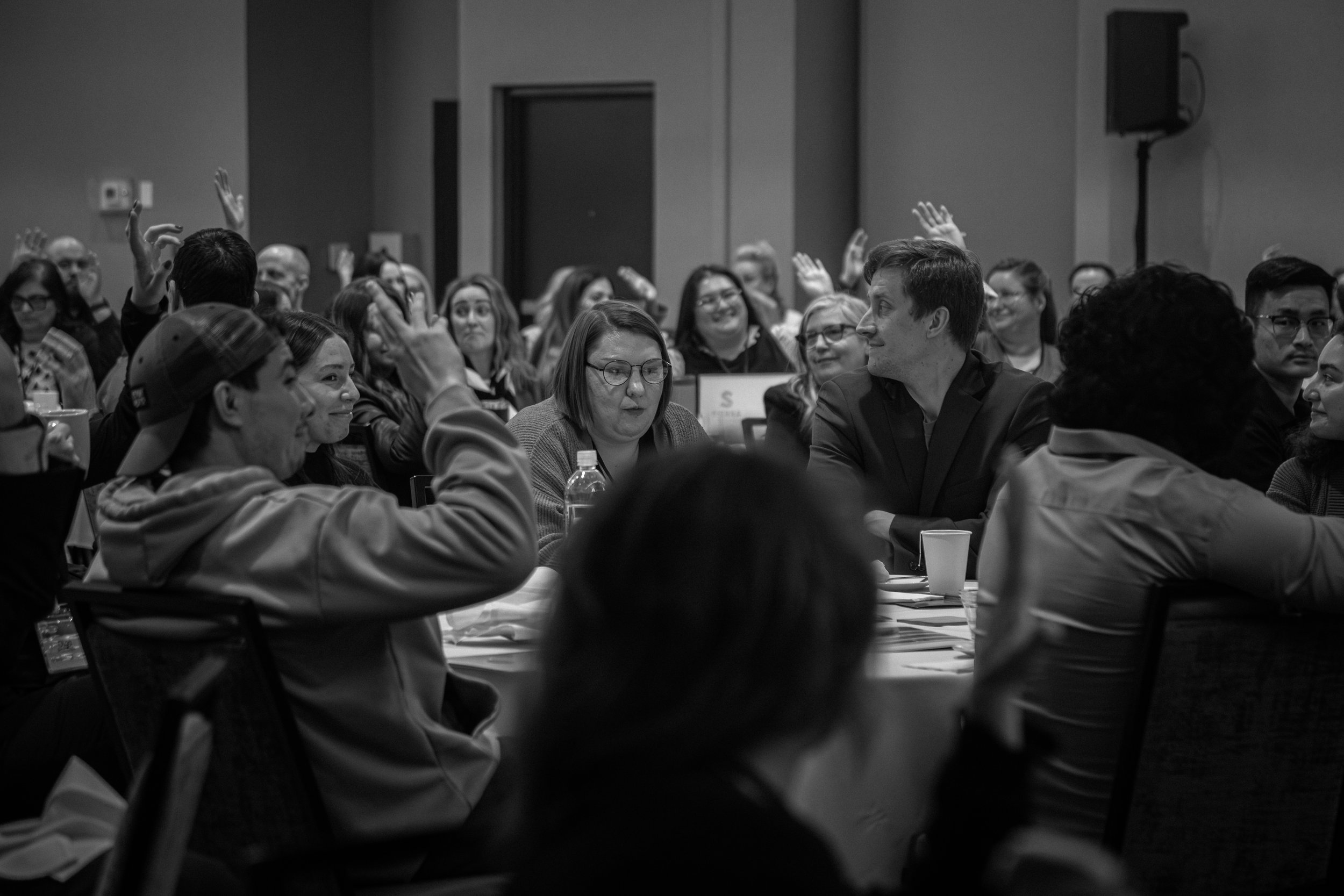 Group of people attending a conference or meeting, with some raising their hands, seated around tables in a large room.