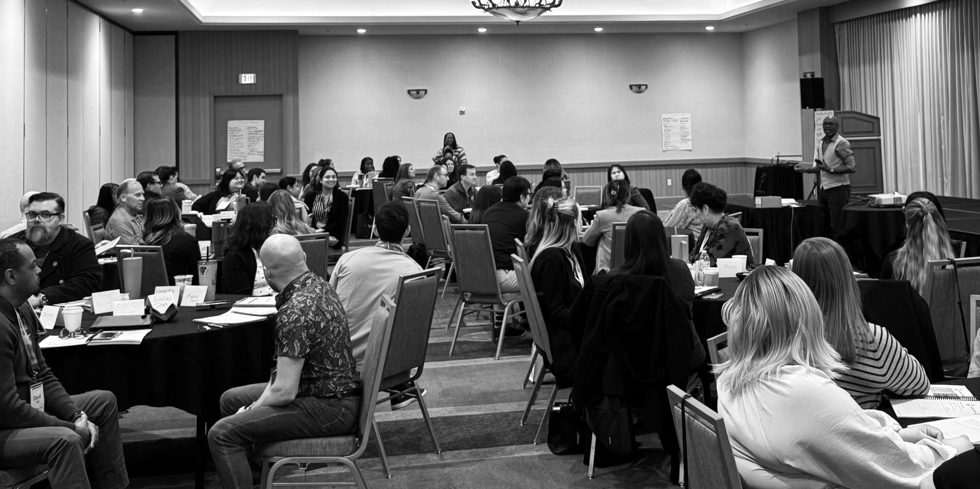 A black and white photo of a large conference room filled with diverse attendees seated at round tables listening to a speaker on a stage.