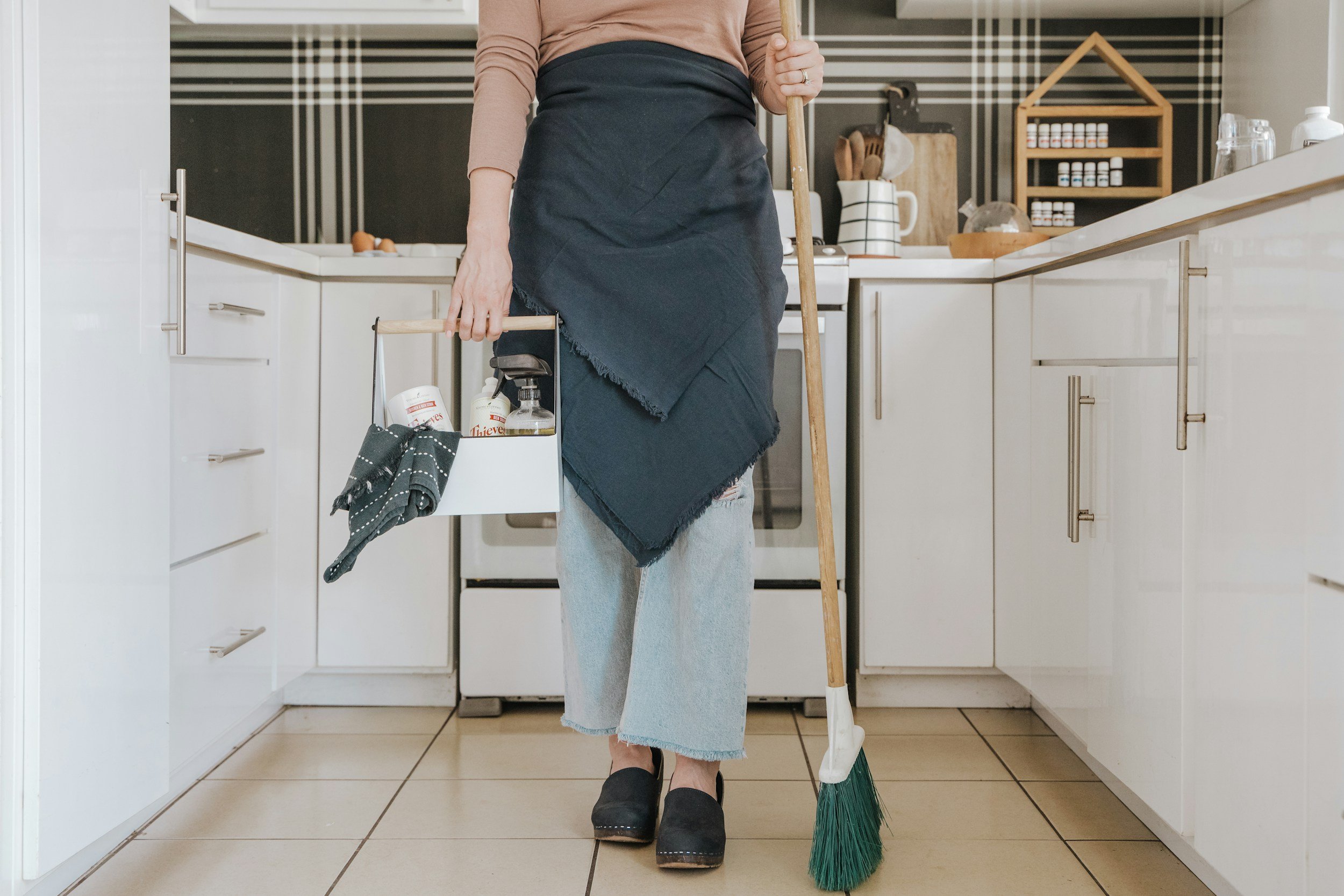 Person cleaning kitchen floor with a broom, wearing jeans, black slippers, and an apron, holding cleaning supplies.