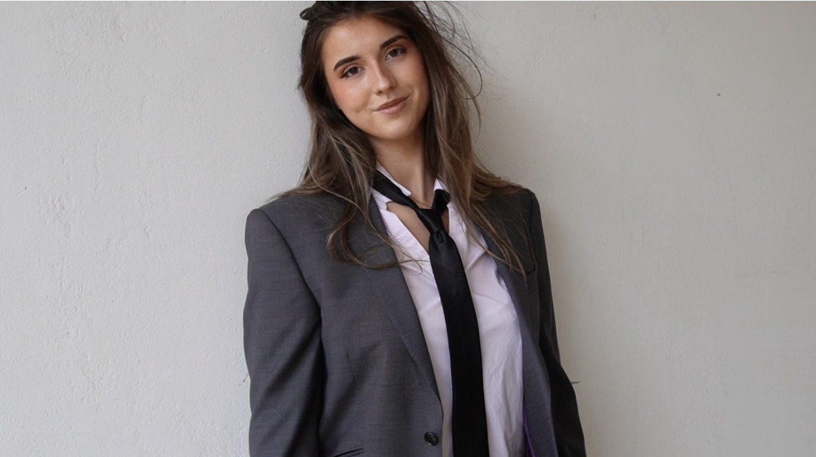 A young woman with long brown hair in a gray business suit, white shirt, and black tie, leaning against a plain white wall.