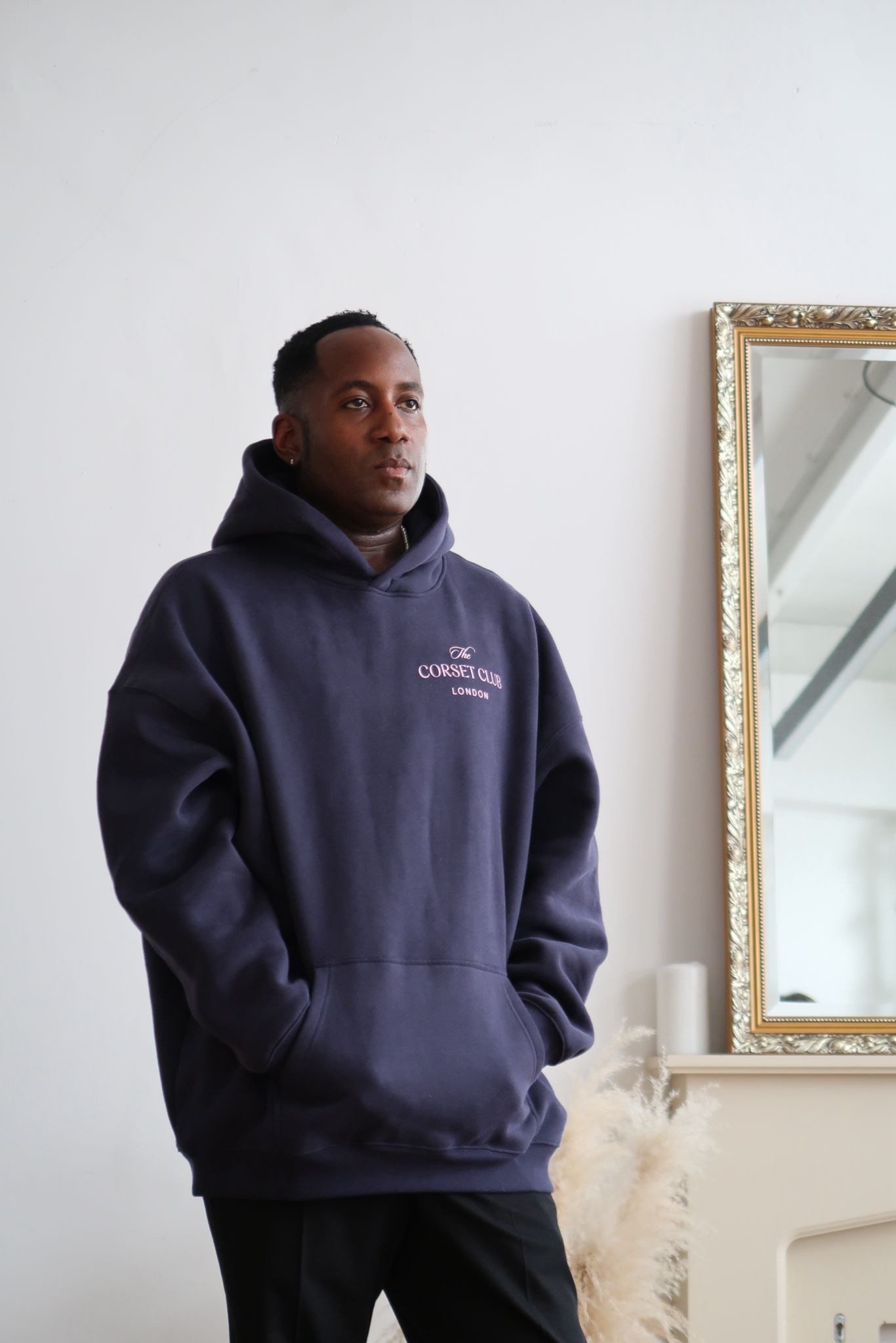A man stands indoors against a white wall, wearing a dark hoodie with 'The Corset Club London' embroidered on it, with hands in pockets, looking slightly to the side, near a large mirror with a gold frame.