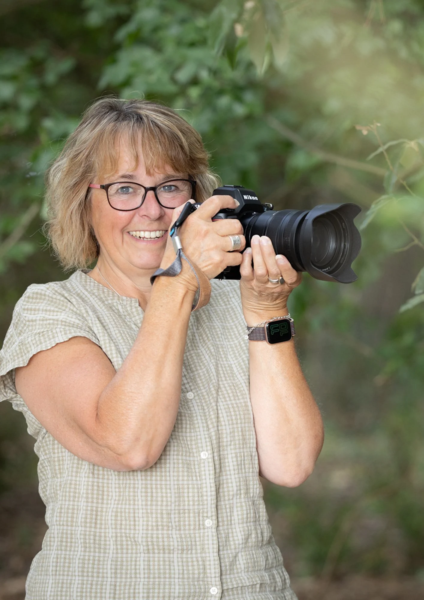 A woman with short, wavy hair, glasses, and a broad smile is holding a camera with a large lens, outdoors surrounded by lush green foliage.