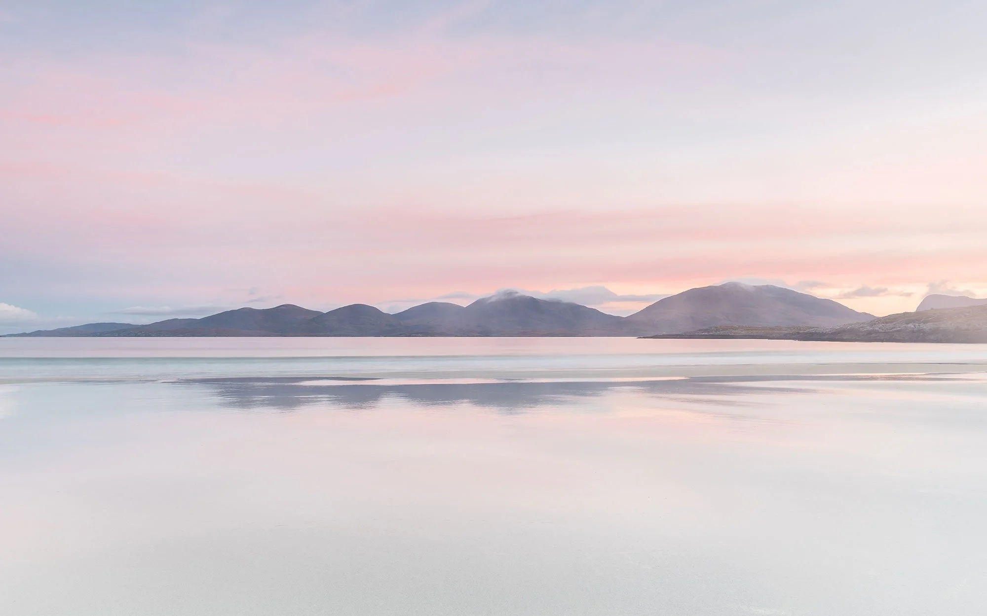 Scenic view of a calm lake with mountains in the background during sunrise, pink and blue sky, and reflections on the water.