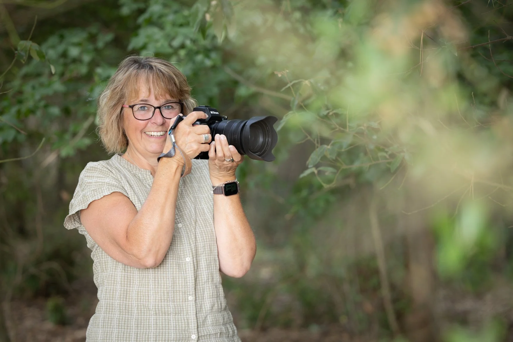 A woman with short light brown hair and glasses, smiling while holding a Nikon camera in a wooded outdoor setting.