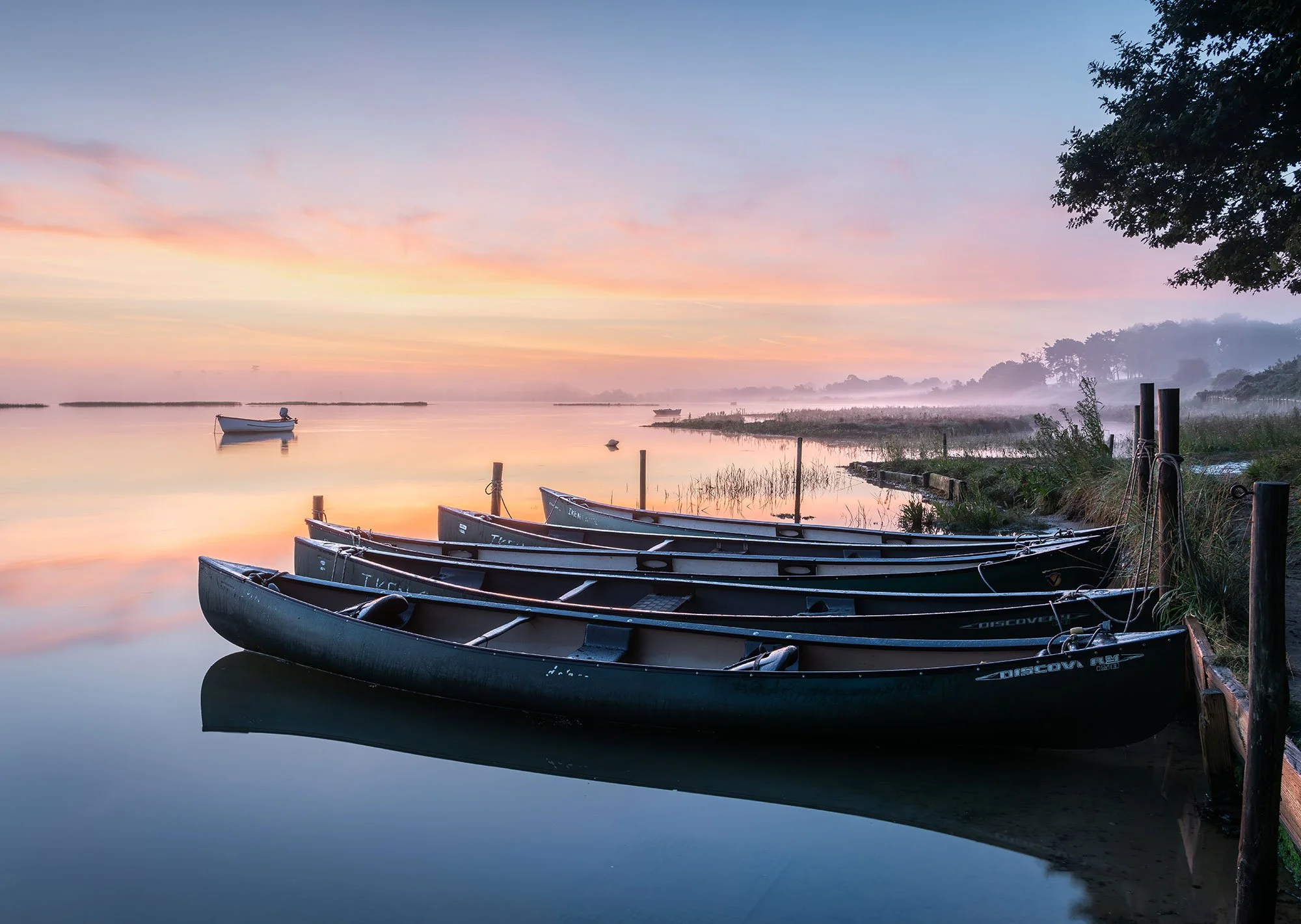 A tranquil lakeside scene at sunrise with several canoes docked along the shore, calm water reflecting the colorful sky, and mist in the distance near trees.