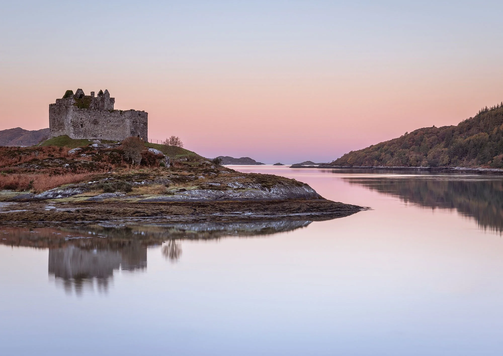 Ancient castle on a hill overlooking a calm body of water with a colorful sky at sunset or sunrise, and a reflection of the castle in the water.