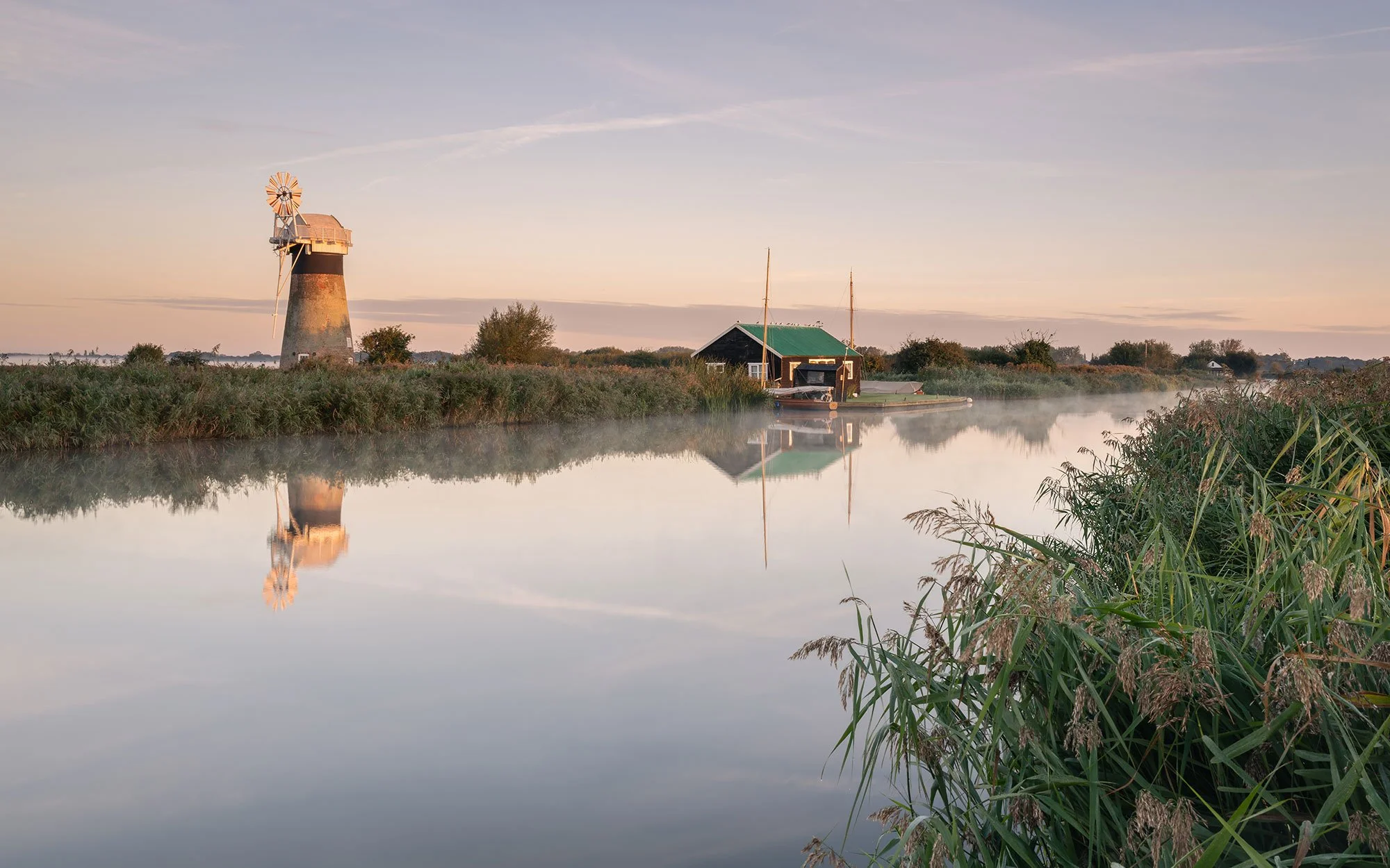A peaceful waterway scene at dawn with a traditional windmill on the left, a small boathouse and sailboat in the center, and tall grasses along the right bank, all reflected in the calm water.