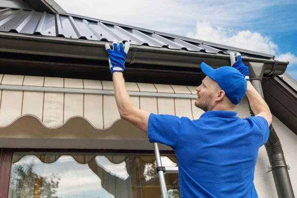 A man wearing a blue uniform and cap, standing on a ladder, installing or repairing a gutter on a house with a metal roof and a window below.