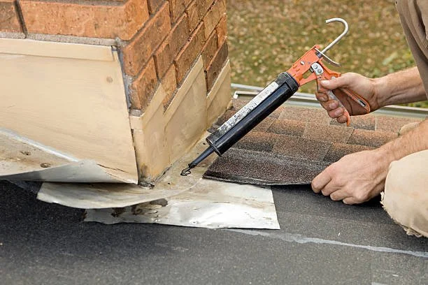 Person using a caulking gun to apply sealant around a chimney on a roof.