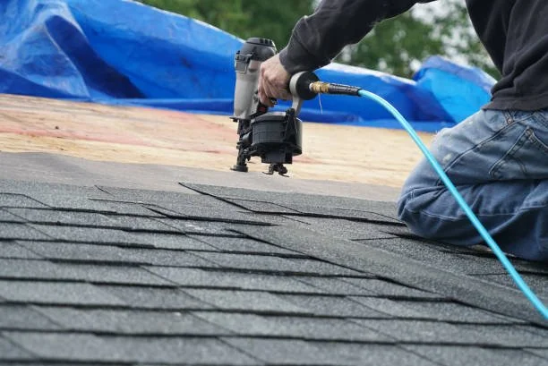 A person kneeling on a roof, using a nail gun connected to an air compressor to install asphalt shingles.