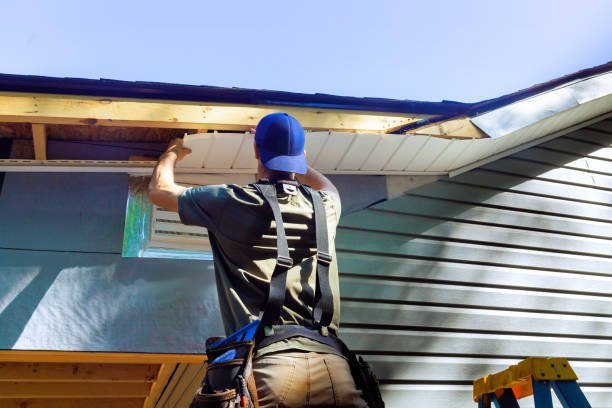 A person wearing a blue cap and work clothes is installing or repairing the roof of a house, using a ladder for support.
