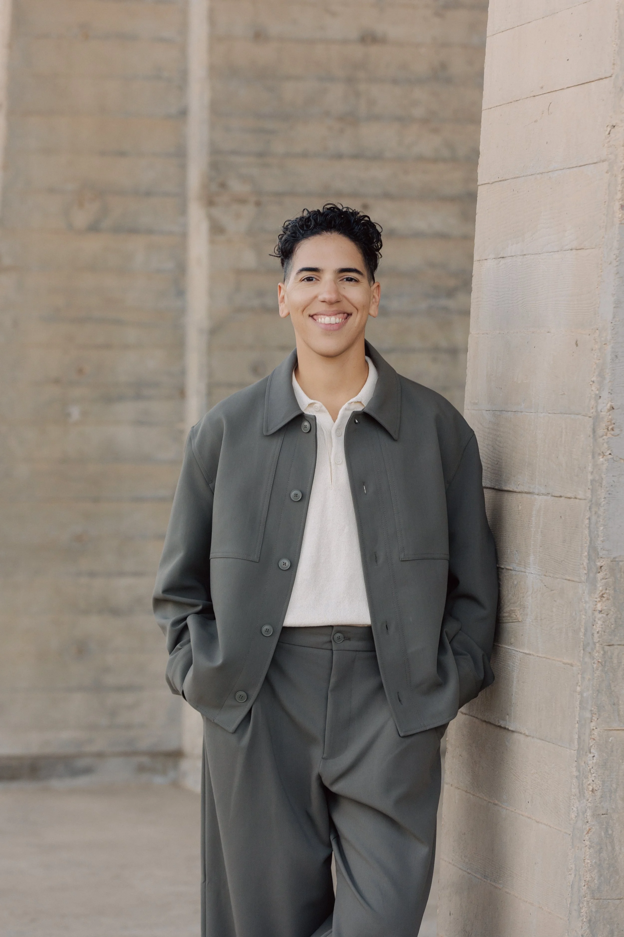 A smiling person with short curly hair leaning against a brick wall in an outdoor setting.