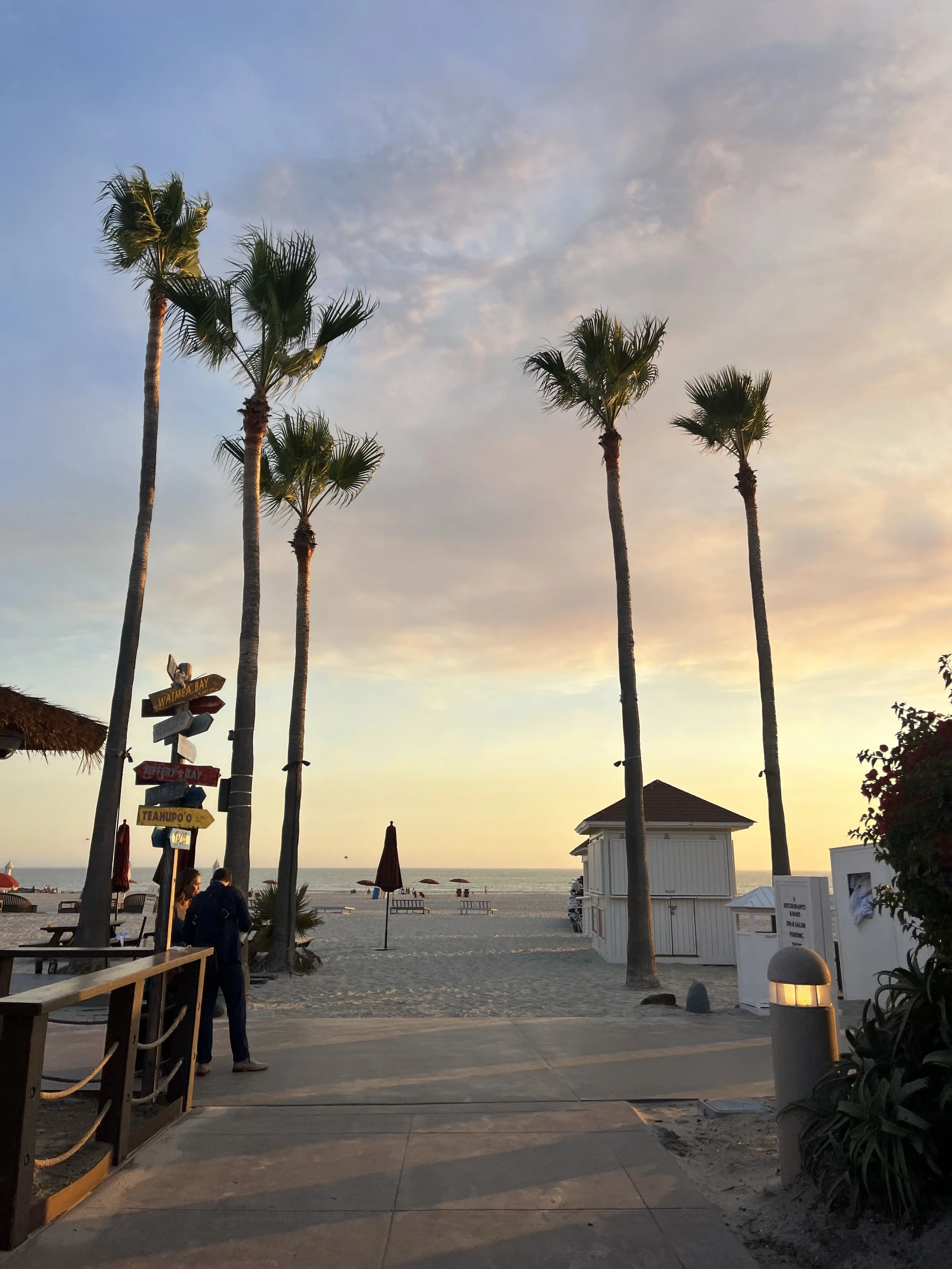 Sunset at a beach with seven tall palm trees, beach umbrellas, lounge chairs, and a small white beach hut. There are a few people standing near the entrance, and the sea is visible in the background with a calm ocean and a partly cloudy sky.