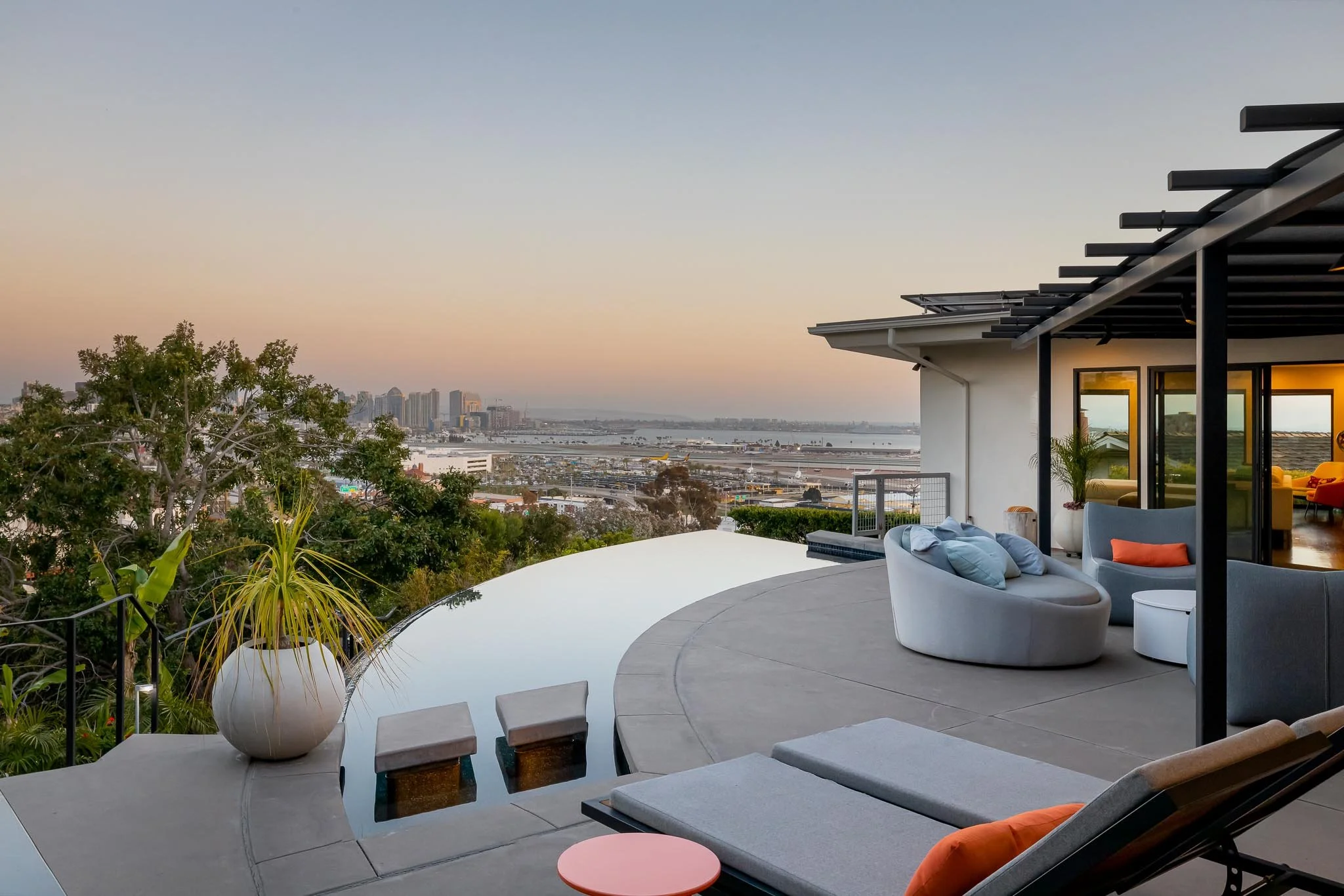 Modern rooftop terrace with outdoor lounge furniture, potted plants, and a view of the city skyline and bay at sunset.