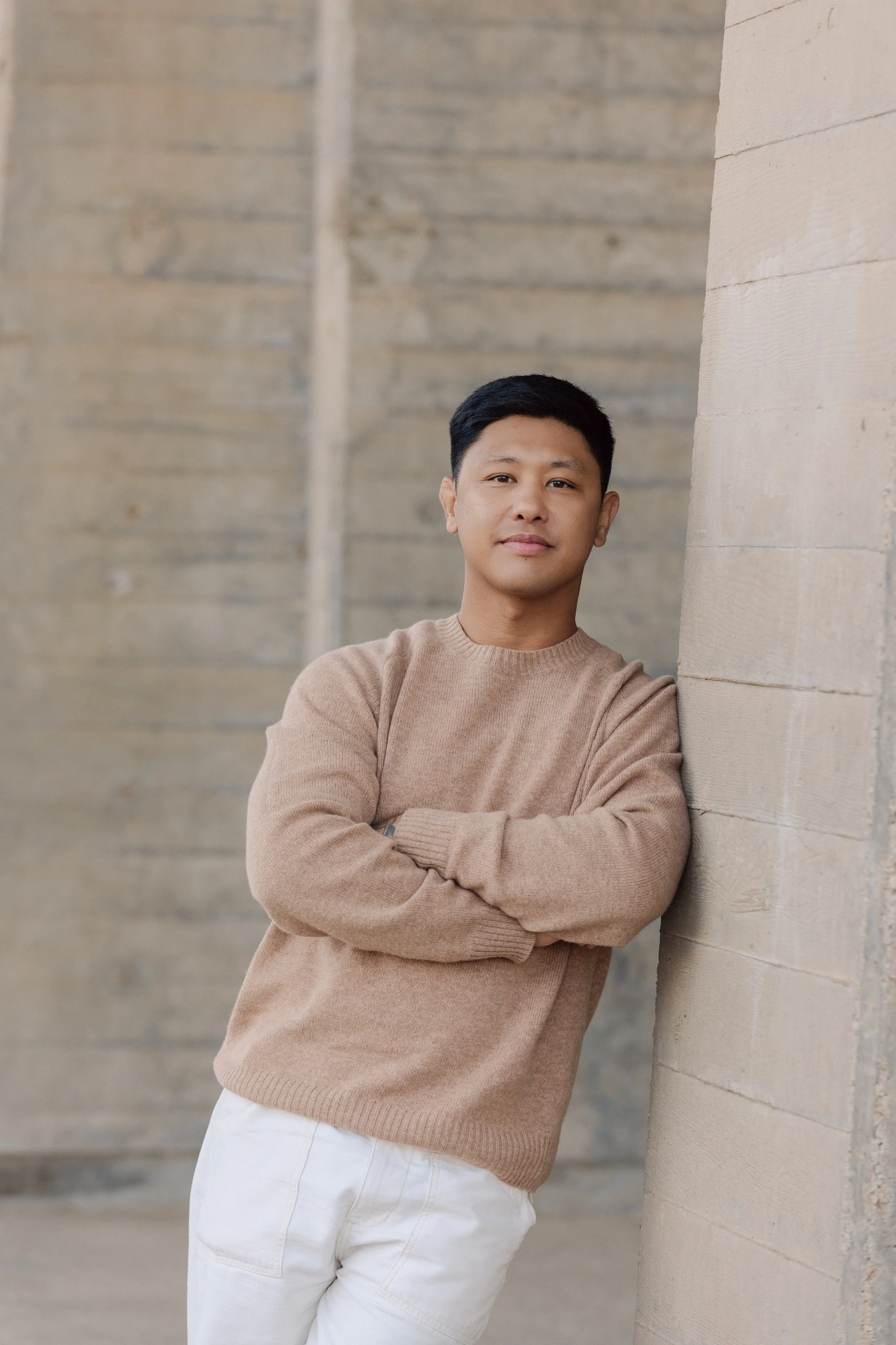 Young man leaning against a wooden wall with arms crossed, wearing a beige sweater and white pants.
