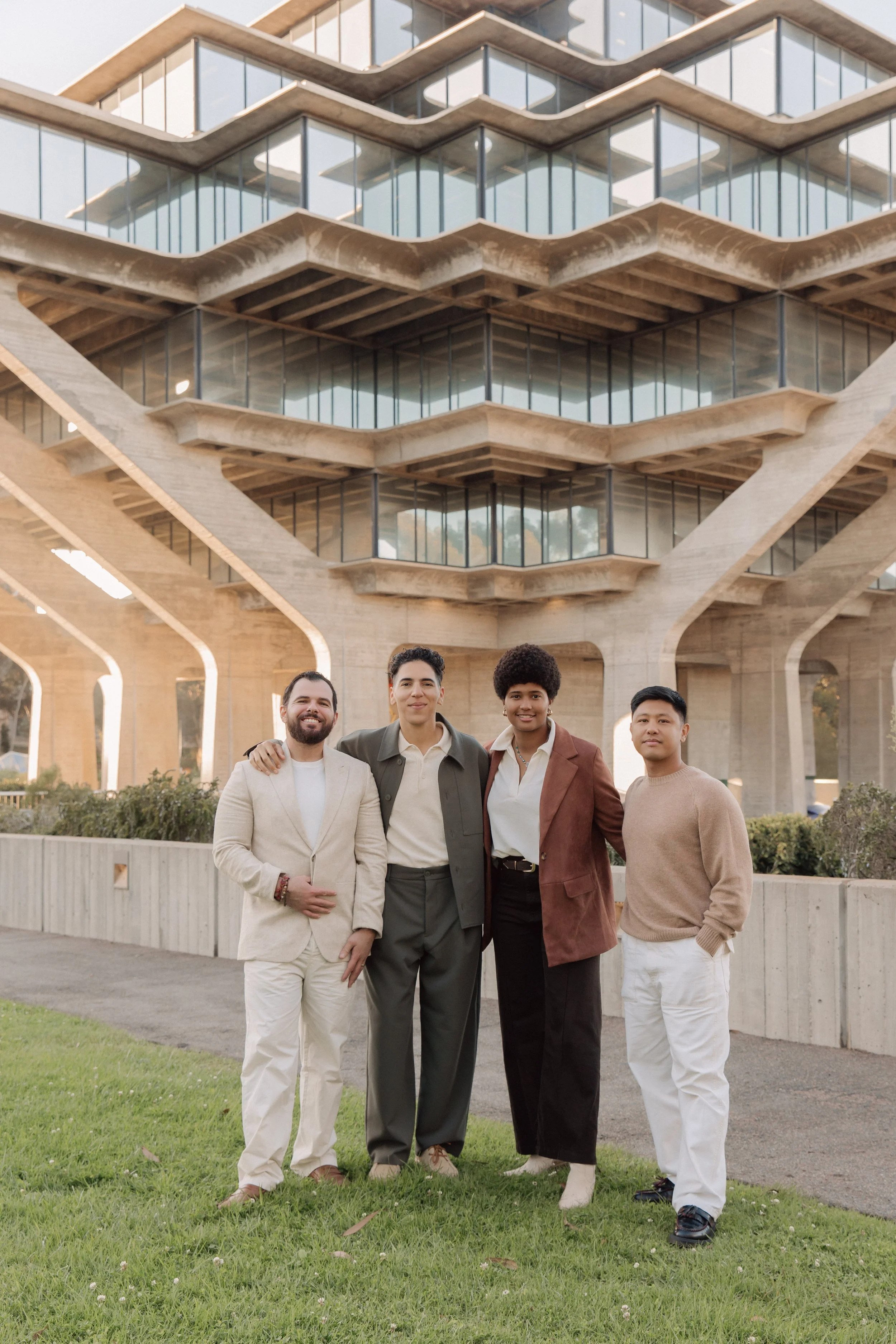Four diverse young adults standing together outdoors in front of a modern architectural building. They are smiling, with arms around each other, dressed in smart casual and semi-formal attire, on a grassy area with some bushes.
